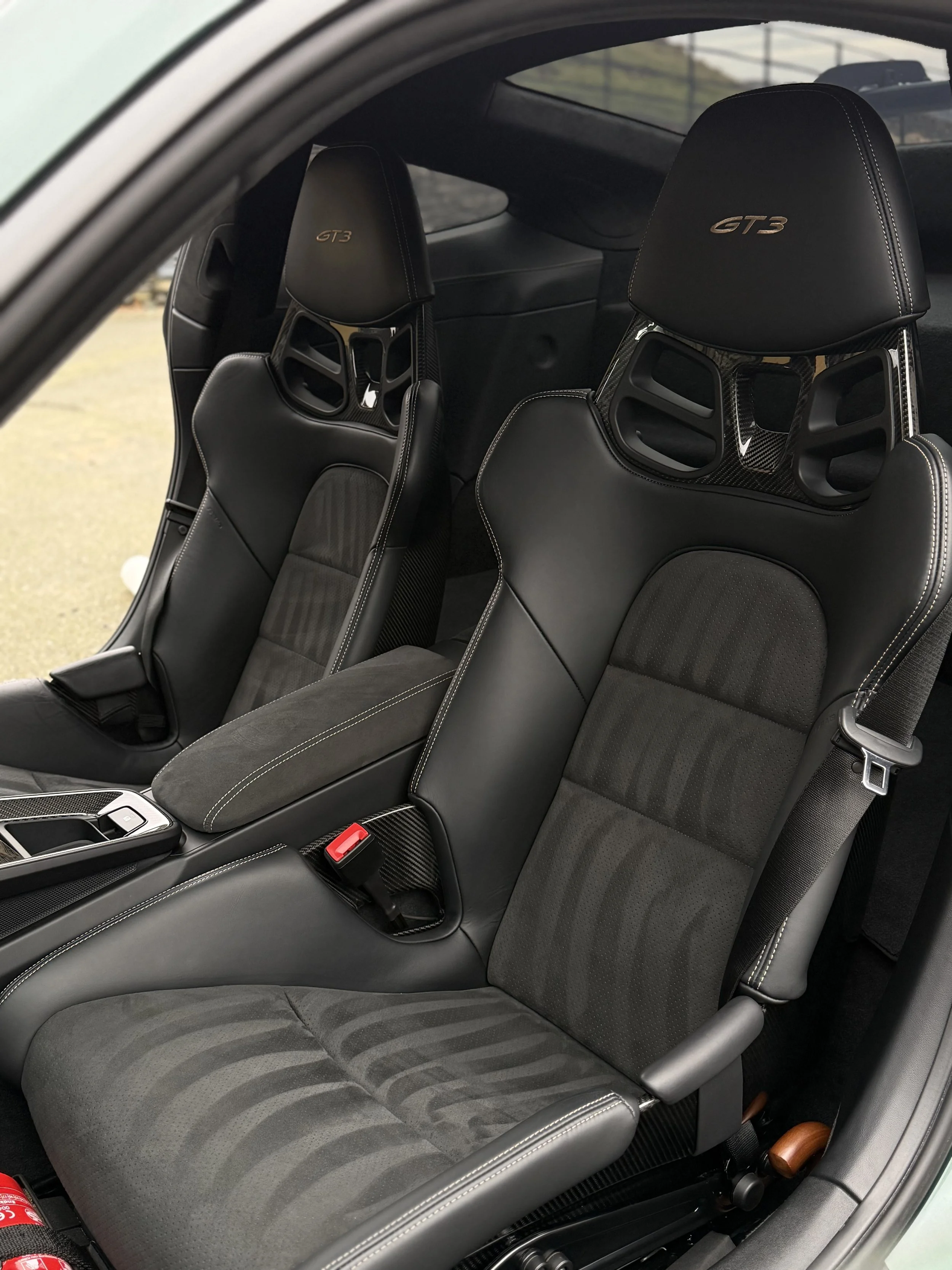 Interior view of a sports car showing black racing seats with the GTX3 logo embroidered on headrests, center console, and seat belts.