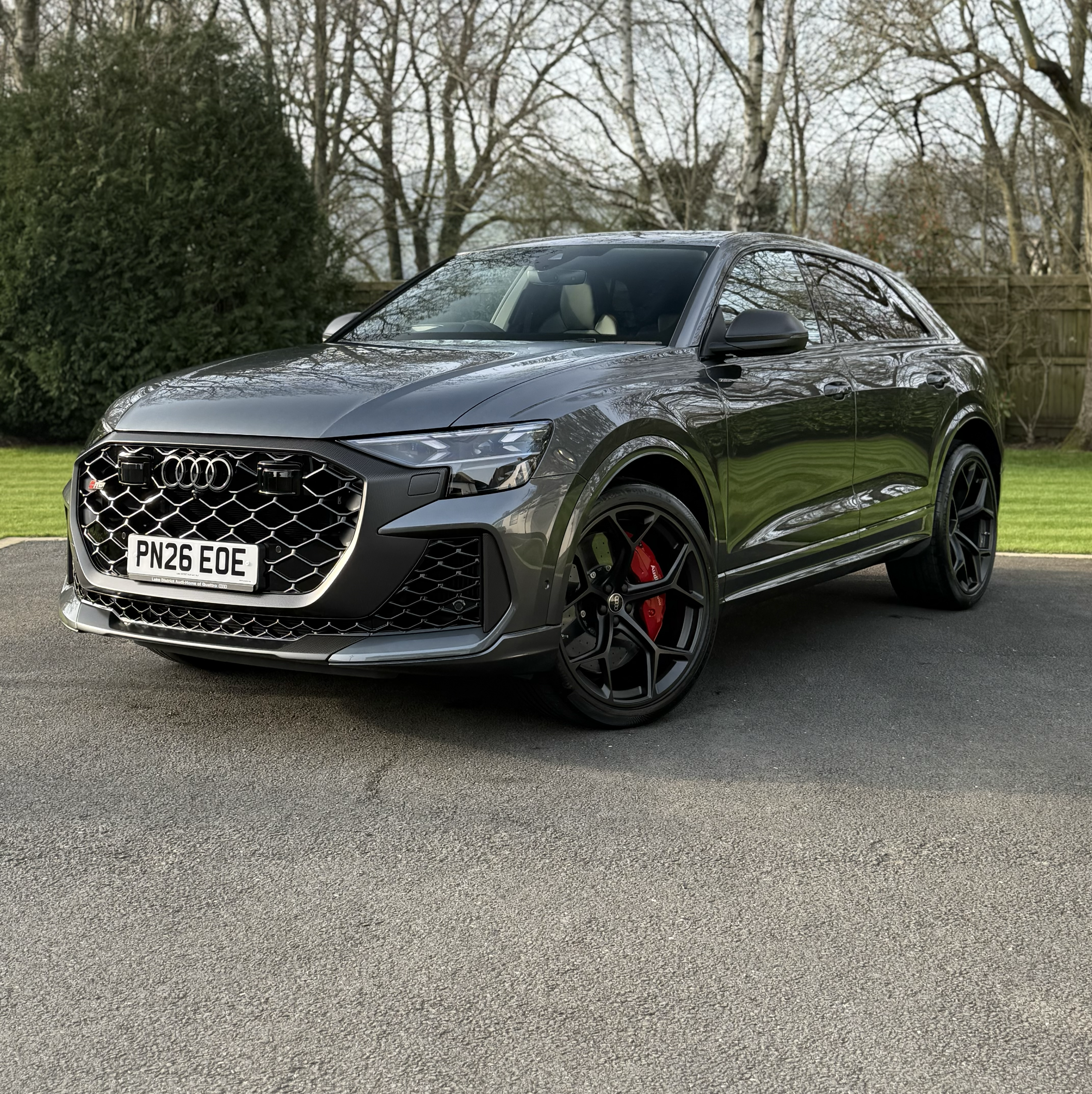 A black Audi hatchback car parked on a driveway with a grassy lawn and a wooden fence in the background, surrounded by leafless trees.