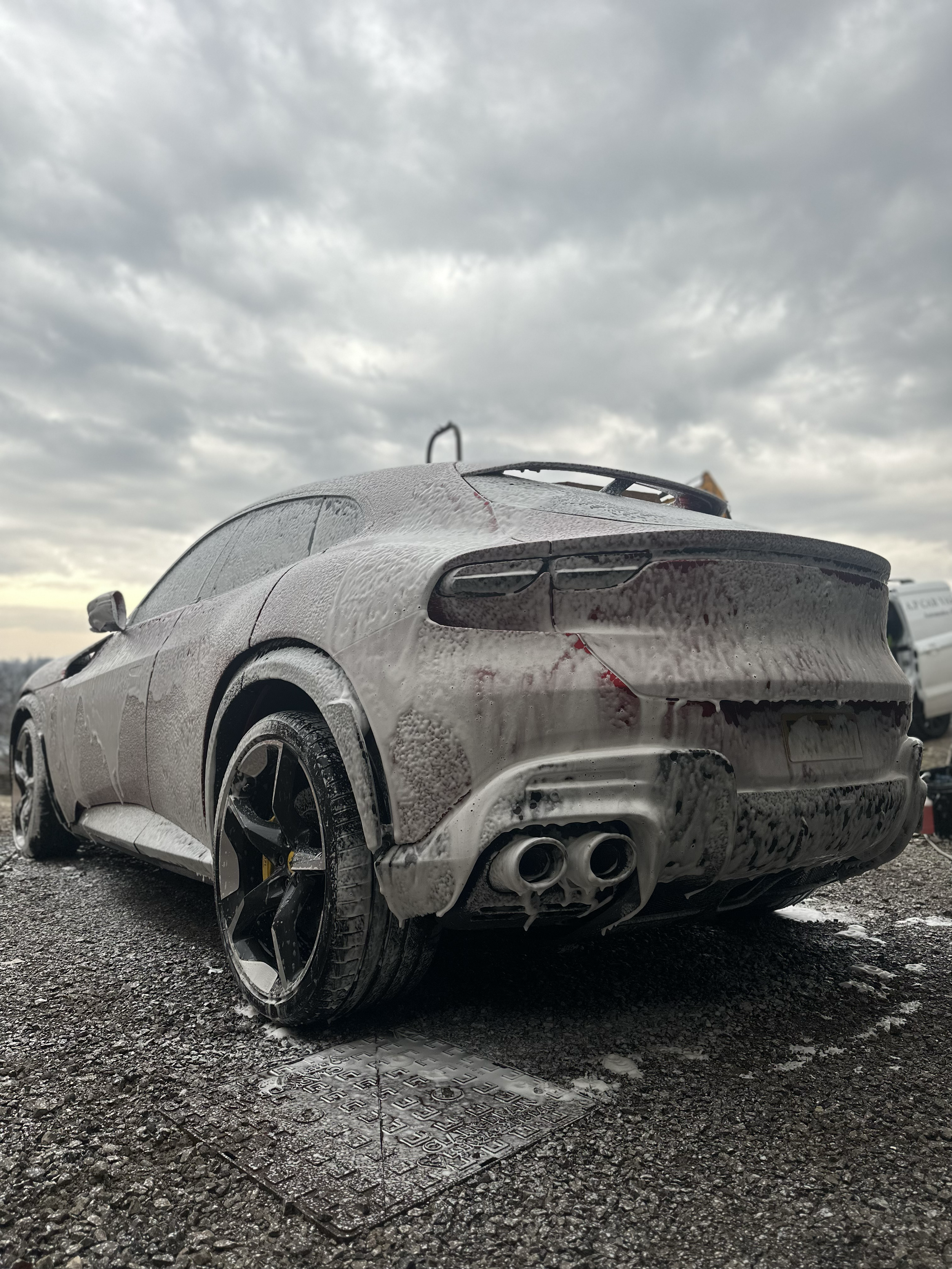 A white sports car covered in foam and soap suds, parked on a wet asphalt surface with a cloudy sky overhead.