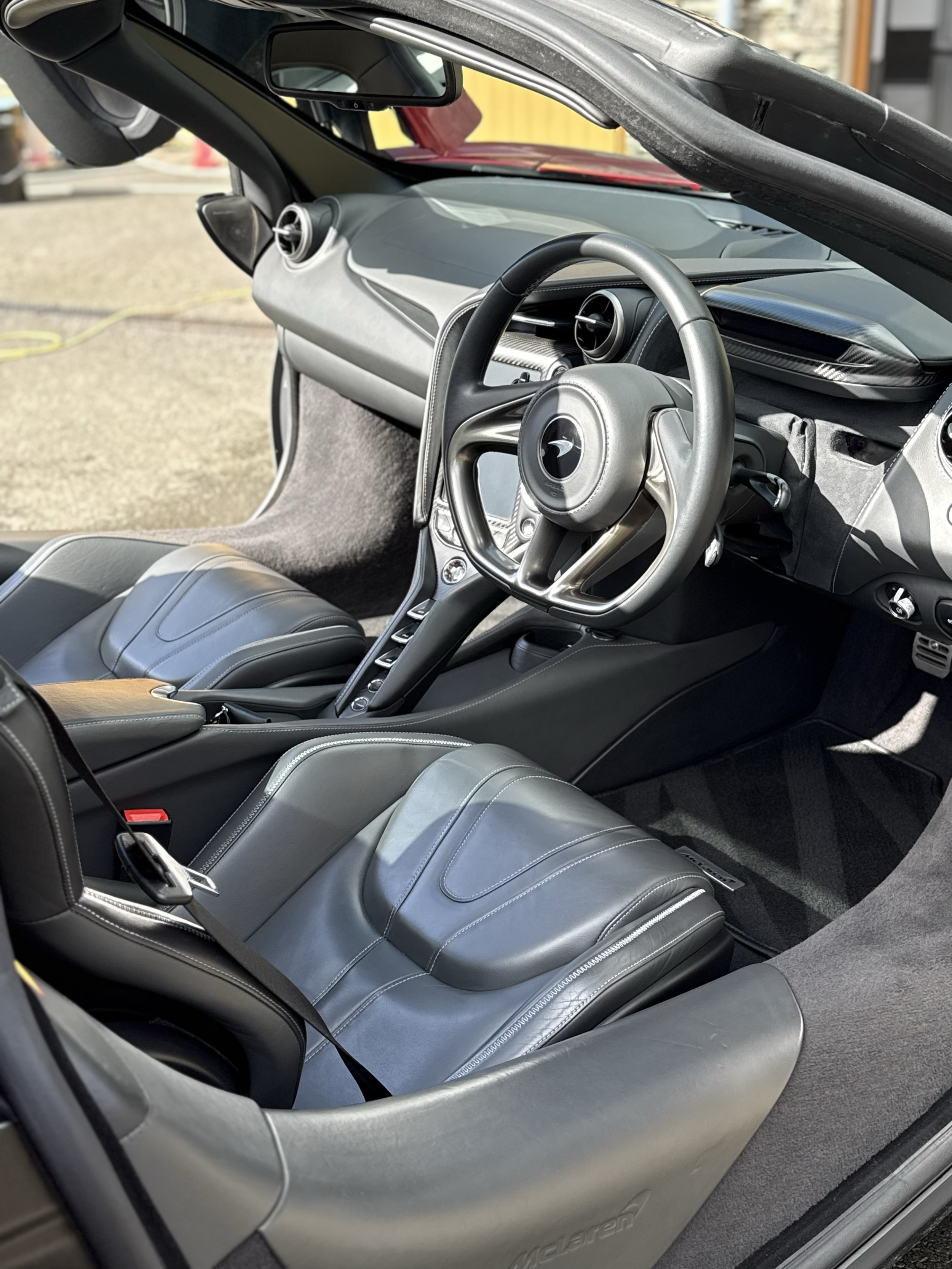 Interior of a sports car with black leather seats, a sleek dashboard, steering wheel, and air vents, parked outdoors.