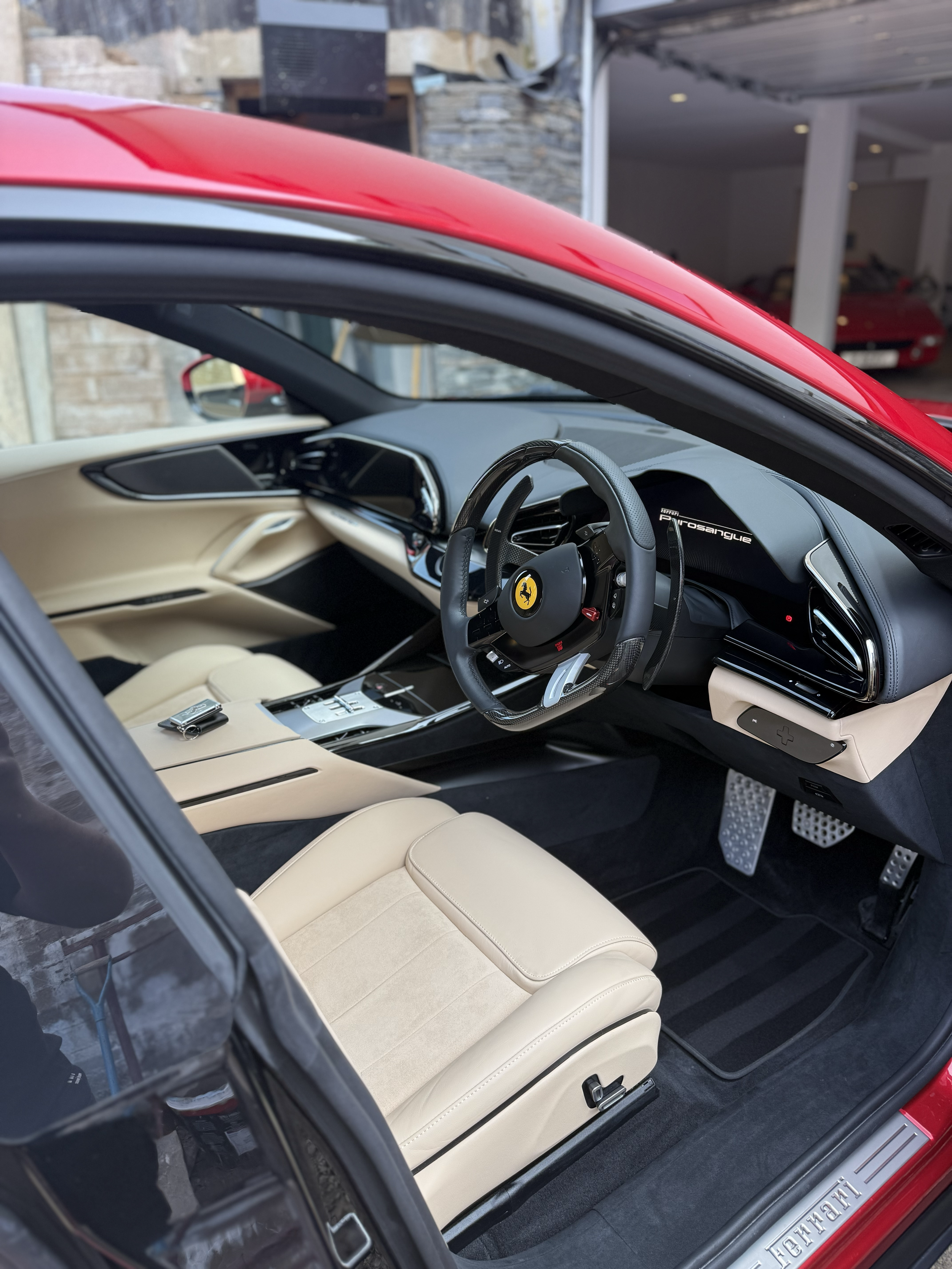 The interior of a luxury red Ferrari sports car, featuring a beige leather seat, a black and leather steering wheel with Ferrari logo, digital dashboard, and metallic pedals.