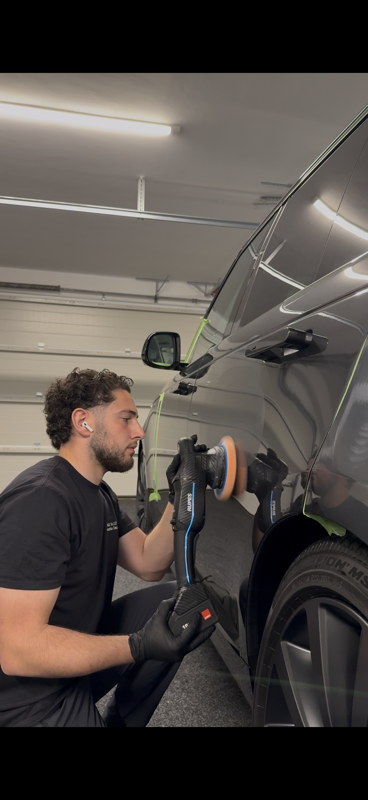 A man is polishing the side of a black SUV with a power buffer, wearing black gloves, black t-shirt, and wireless earbuds in a garage.