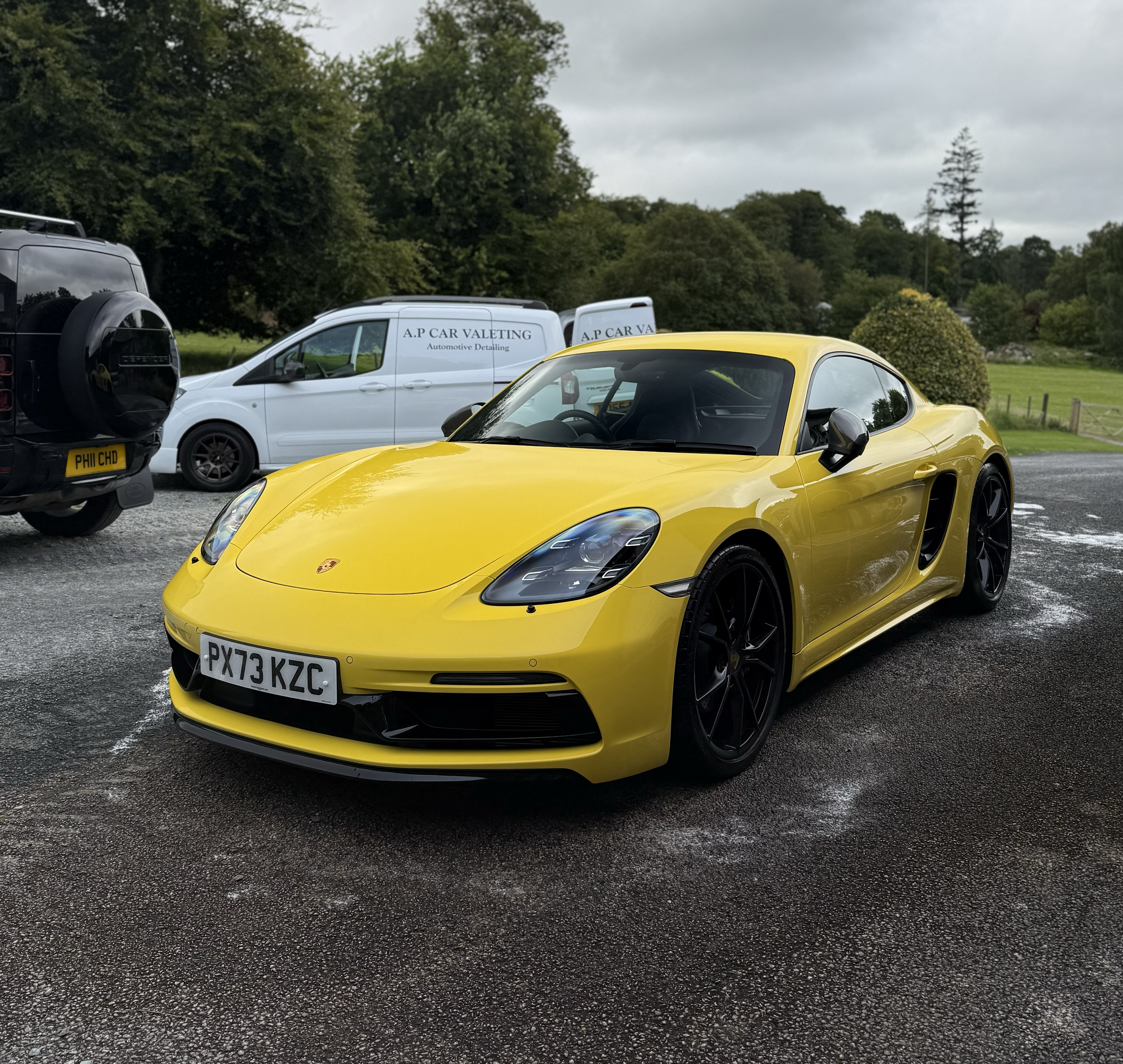 A yellow sports car parked in a parking lot with other vehicles and a background of trees and cloudy sky.