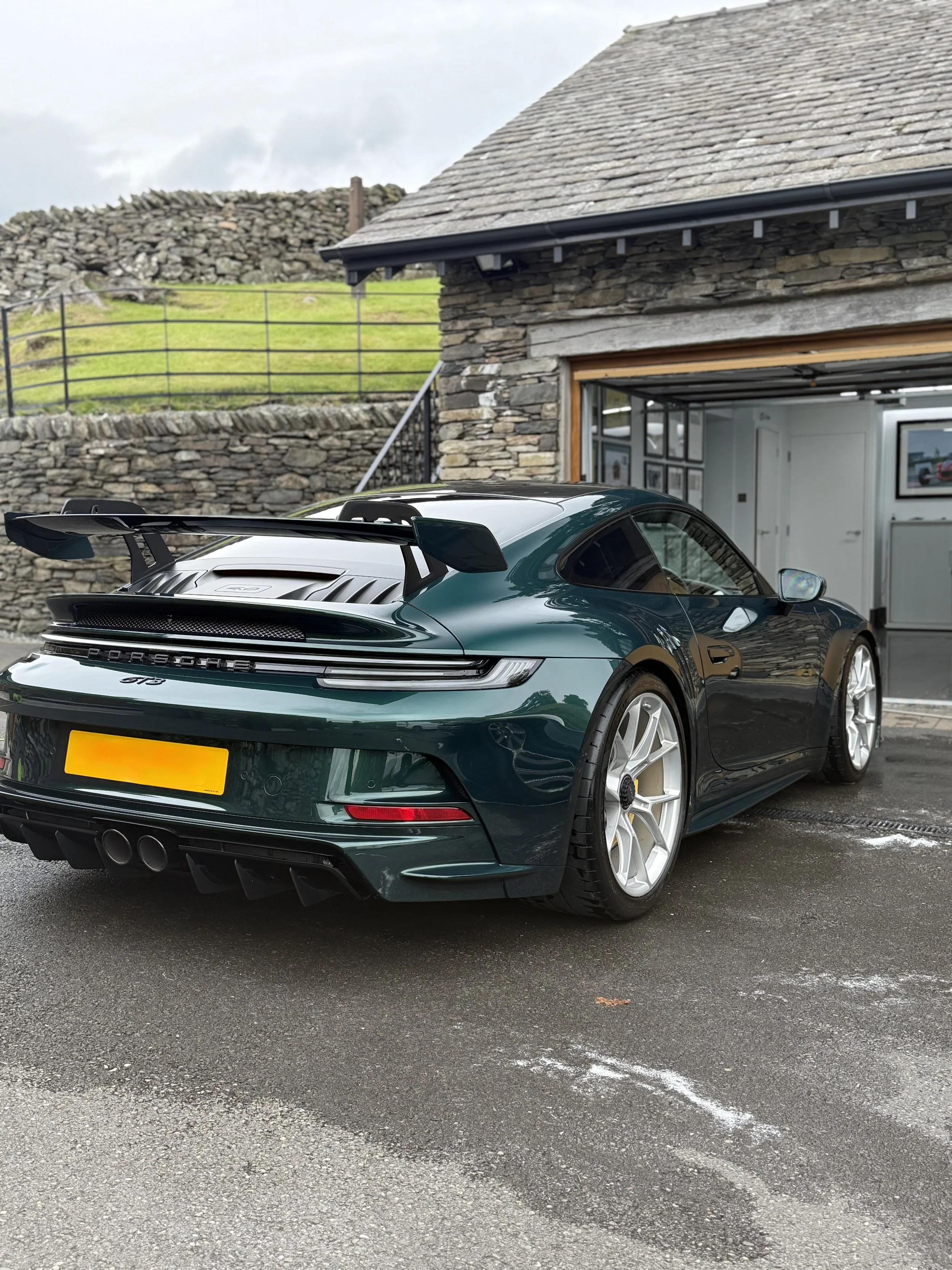 Dark green Porsche 911 GT3 car parked outside a stone building with an open garage door, displaying framed pictures inside, in a setting with a stone wall, railing, and a grassy hill in the background.