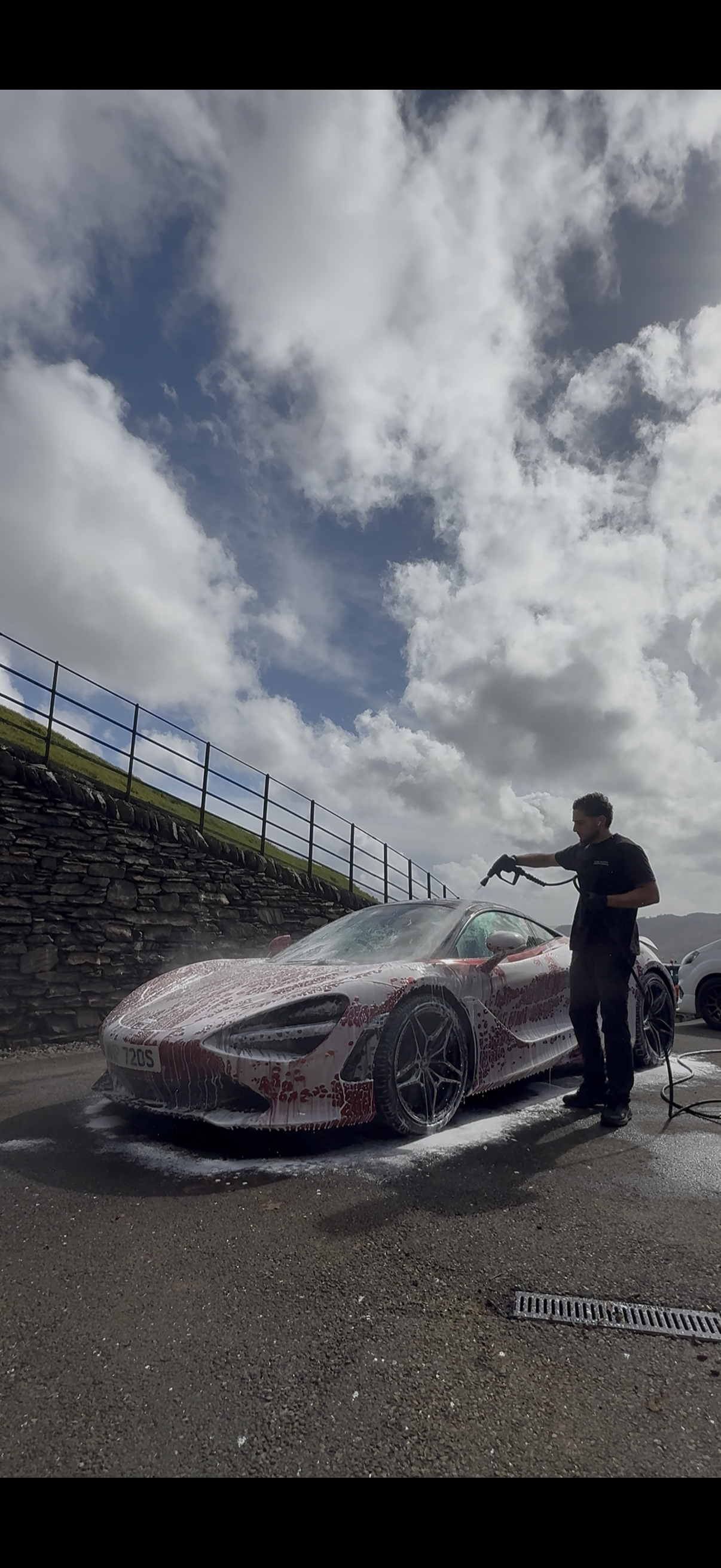A man washing a red sports car with soap suds in an outdoor parking lot, under a partly cloudy sky.