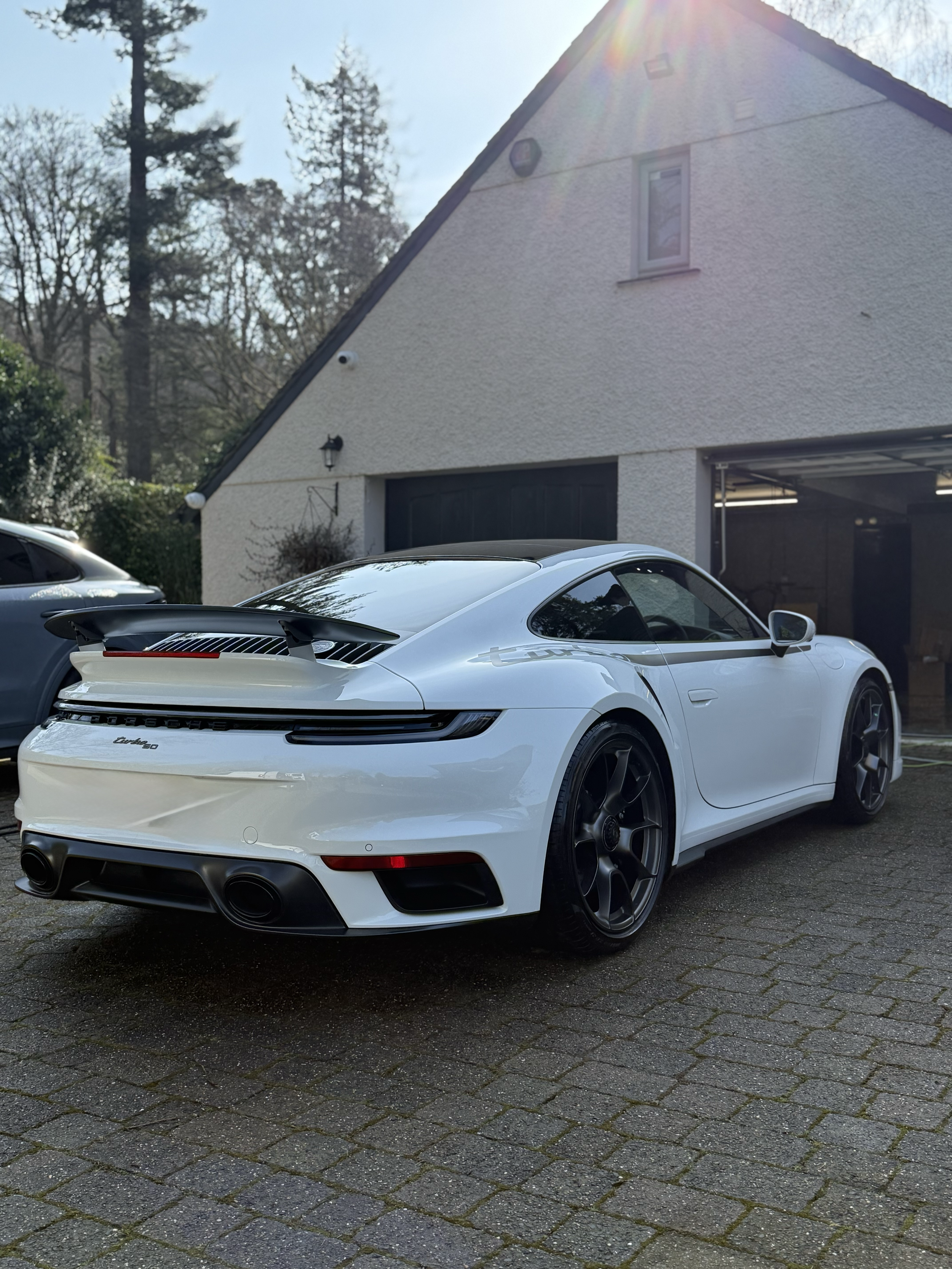 A white sports car parked in front of a house garage, with trees and clear sky in the background, sunlight shining down.