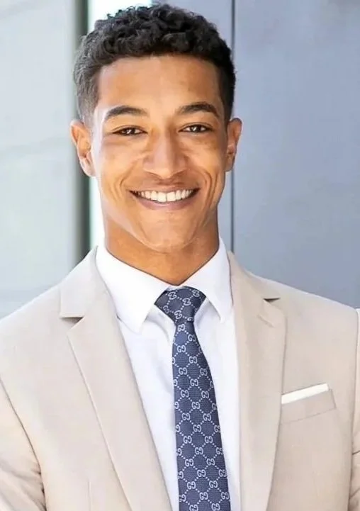 A young man with curly black hair, smiling, wearing a beige suit, white shirt, and navy blue tie with a pattern. He is in an office setting with a gray wall in the background.