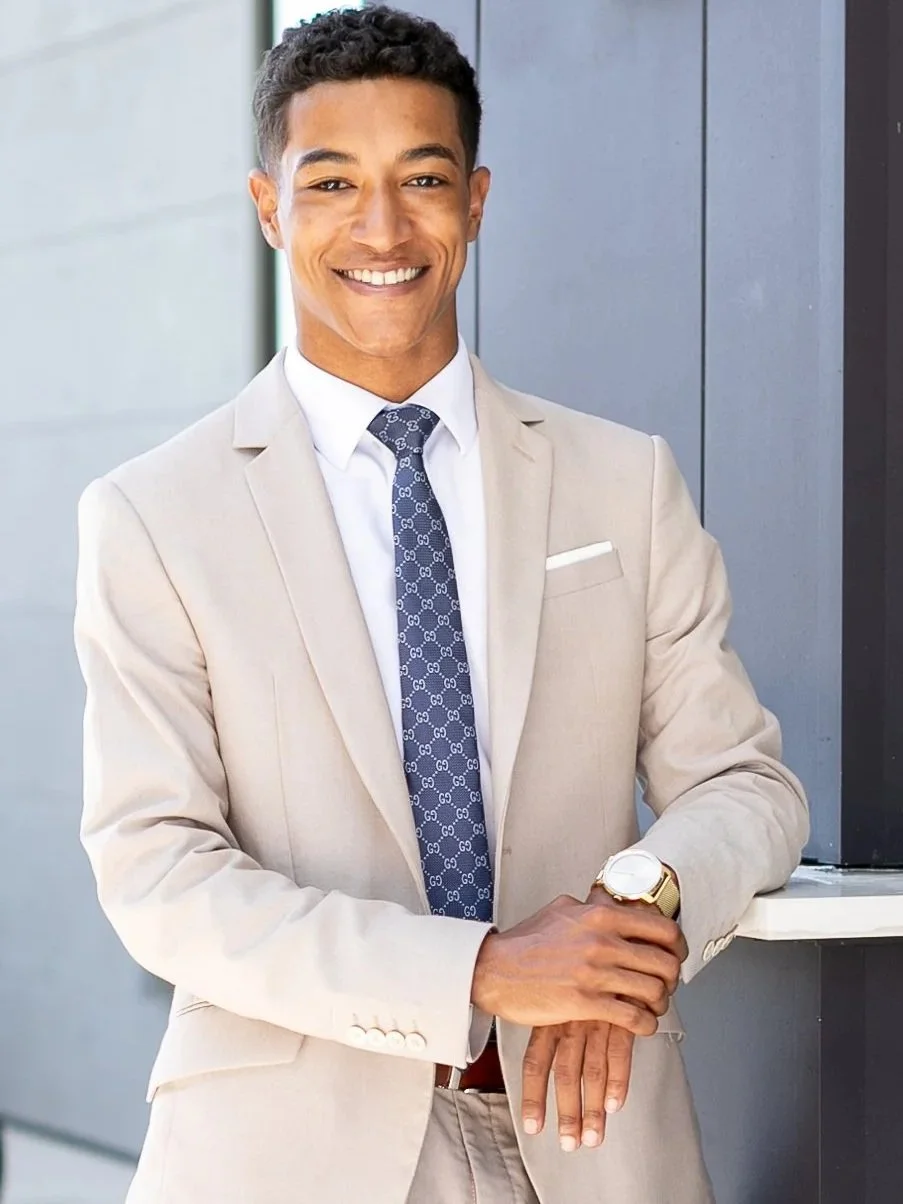 A smartly dressed man in a beige suit, white shirt, and patterned dark blue tie is smiling and leaning on a white surface in an urban setting.