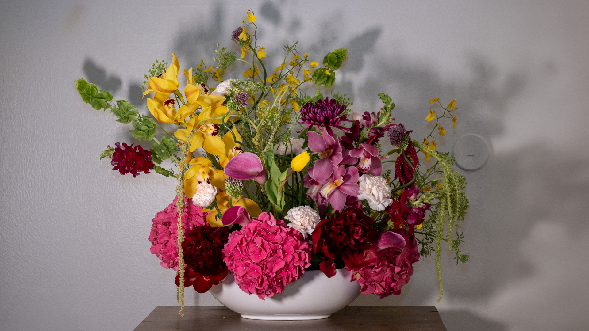 A large, colorful flower arrangement in a round white vase on a wooden table, with a plain light-colored wall in the background.