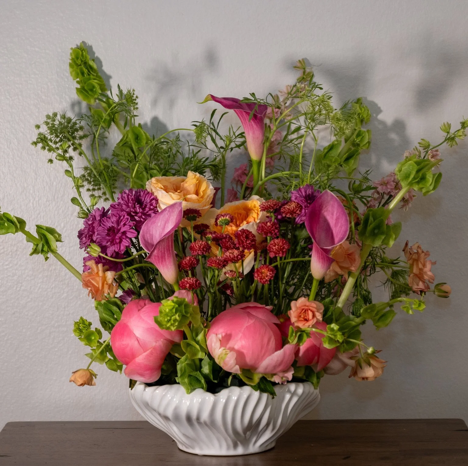 A bouquet of various colorful flowers in a white ceramic vase on a wooden surface against a plain wall.