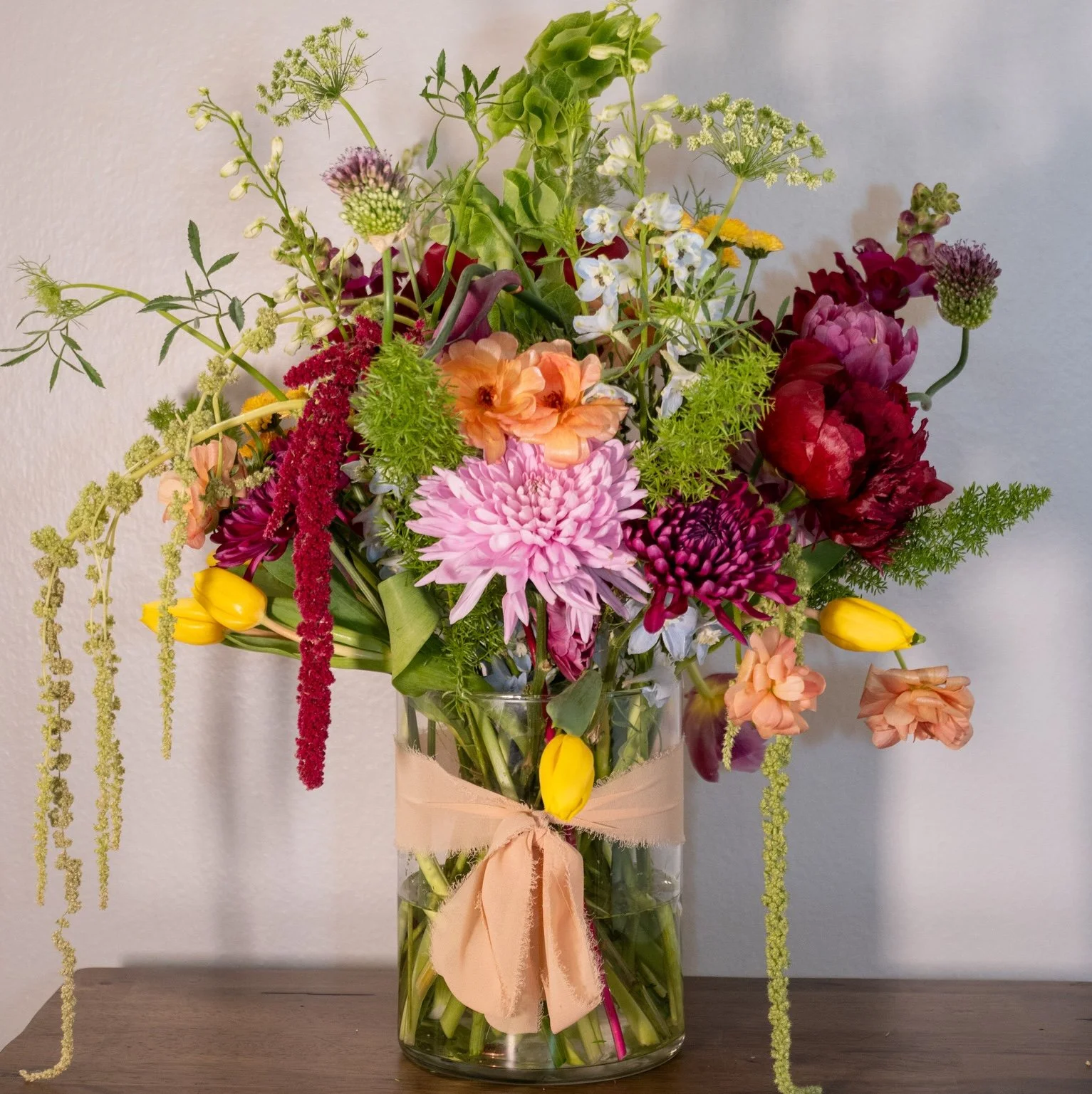 A tall glass vase with a beige ribbon tied around it, filled with a colorful mixed flower bouquet including pink, peach, purple, white, yellow, and green flowers, set on a wooden surface against a light gray wall.