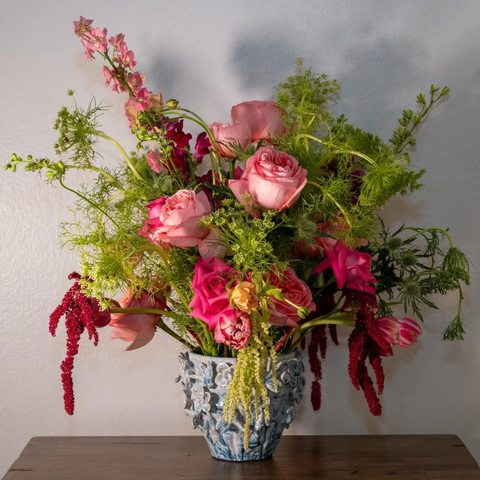 A colorful floral arrangement in a decorative blue and white vase on a wooden table, including pink roses, purple flowers, green foliage, and red hanging amaranthus.