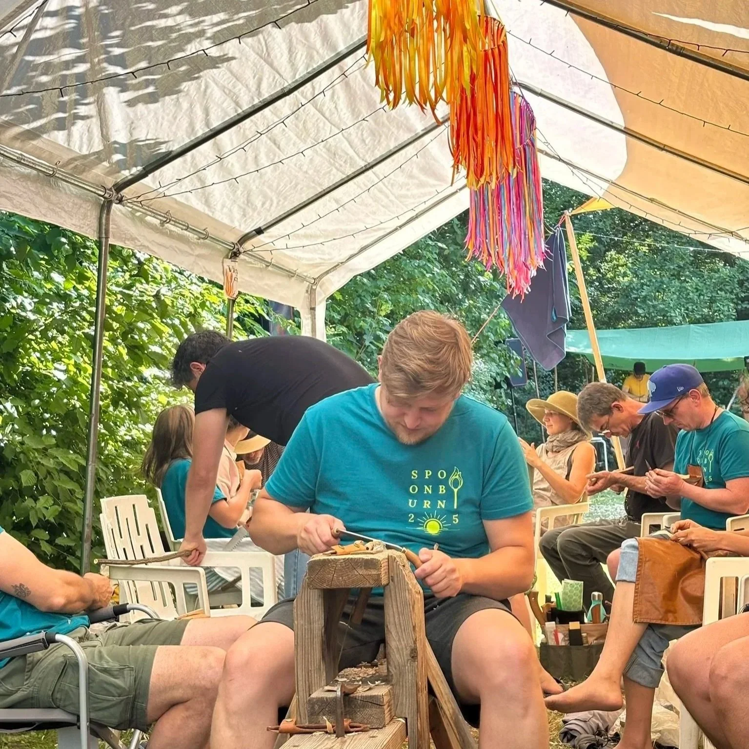 Mensen die getalenteerd houtbewerking doen onder een witte tent, met kleurrijke kunstwerken onder de tent en groene bomen op de achtergrond.