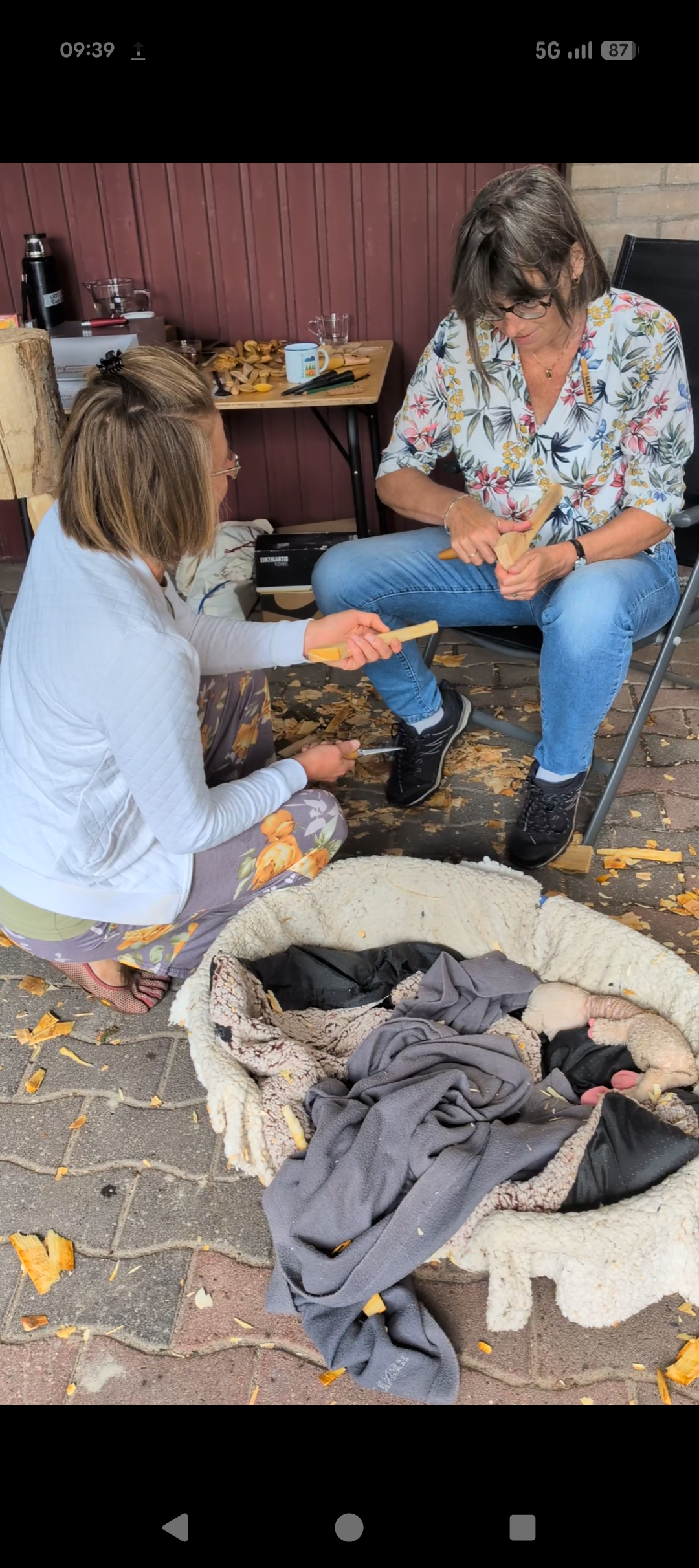 Twee vrouwen hamers en zagen gebruiken om hout te bewerken, zittend op een houten bank buiten, met een mand vol kleding in de buurt.