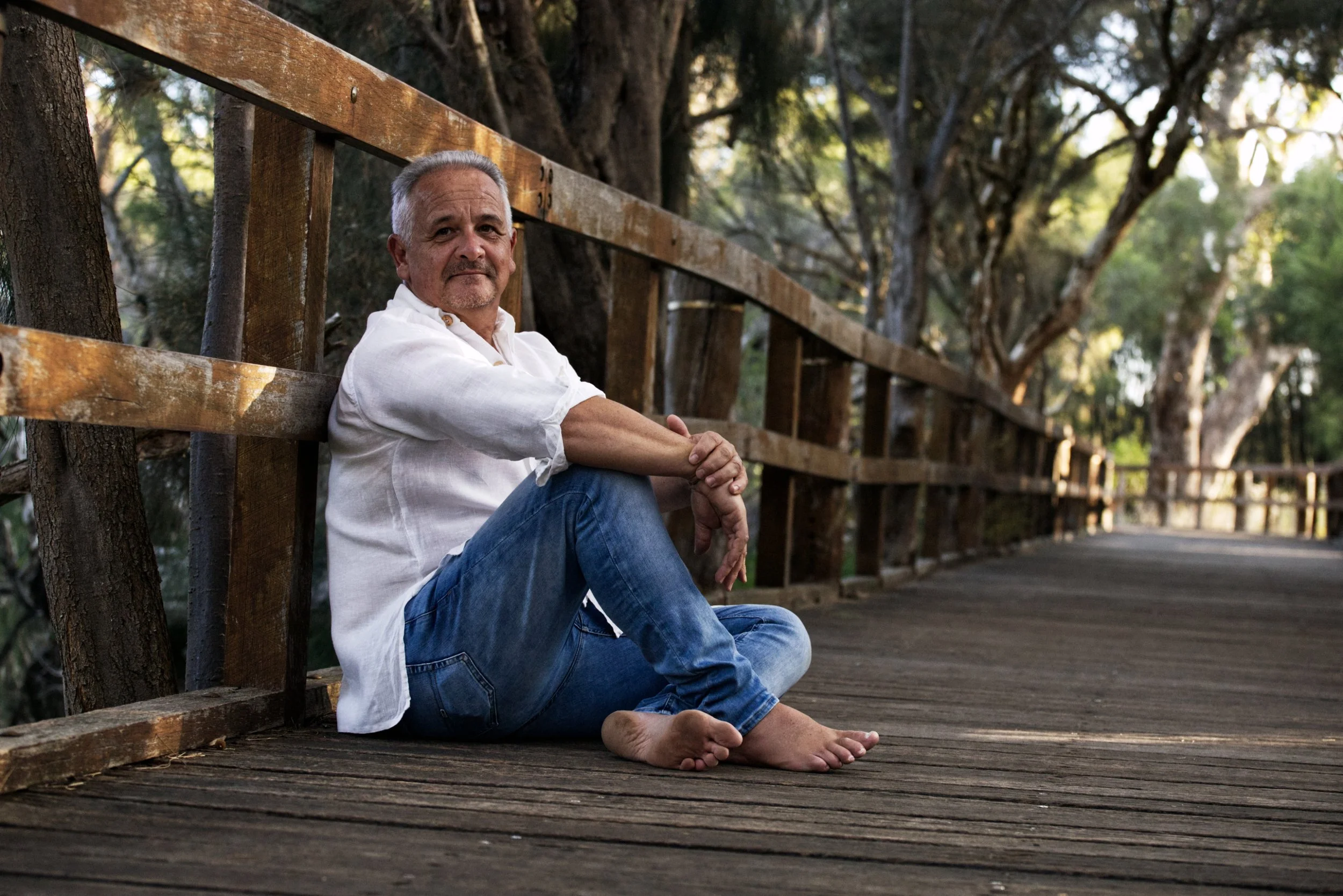 A middle-aged man with gray hair and a beard sits on a wooden boardwalk by a wooden railing, surrounded by large trees with sunlight filtering through the leaves.