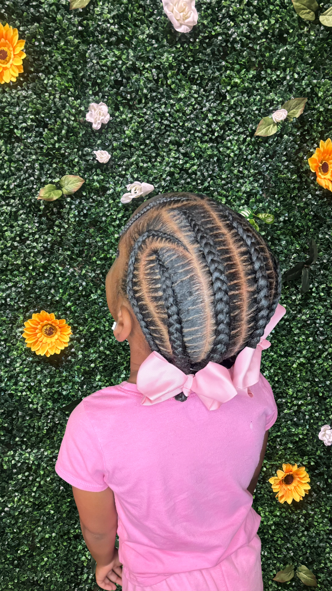 A young girl with intricately braided hair adorned with a pink bow, seen from behind, wearing a pink shirt, standing in front of a green leafy wall decorated with sunflowers, white, and purple flowers.