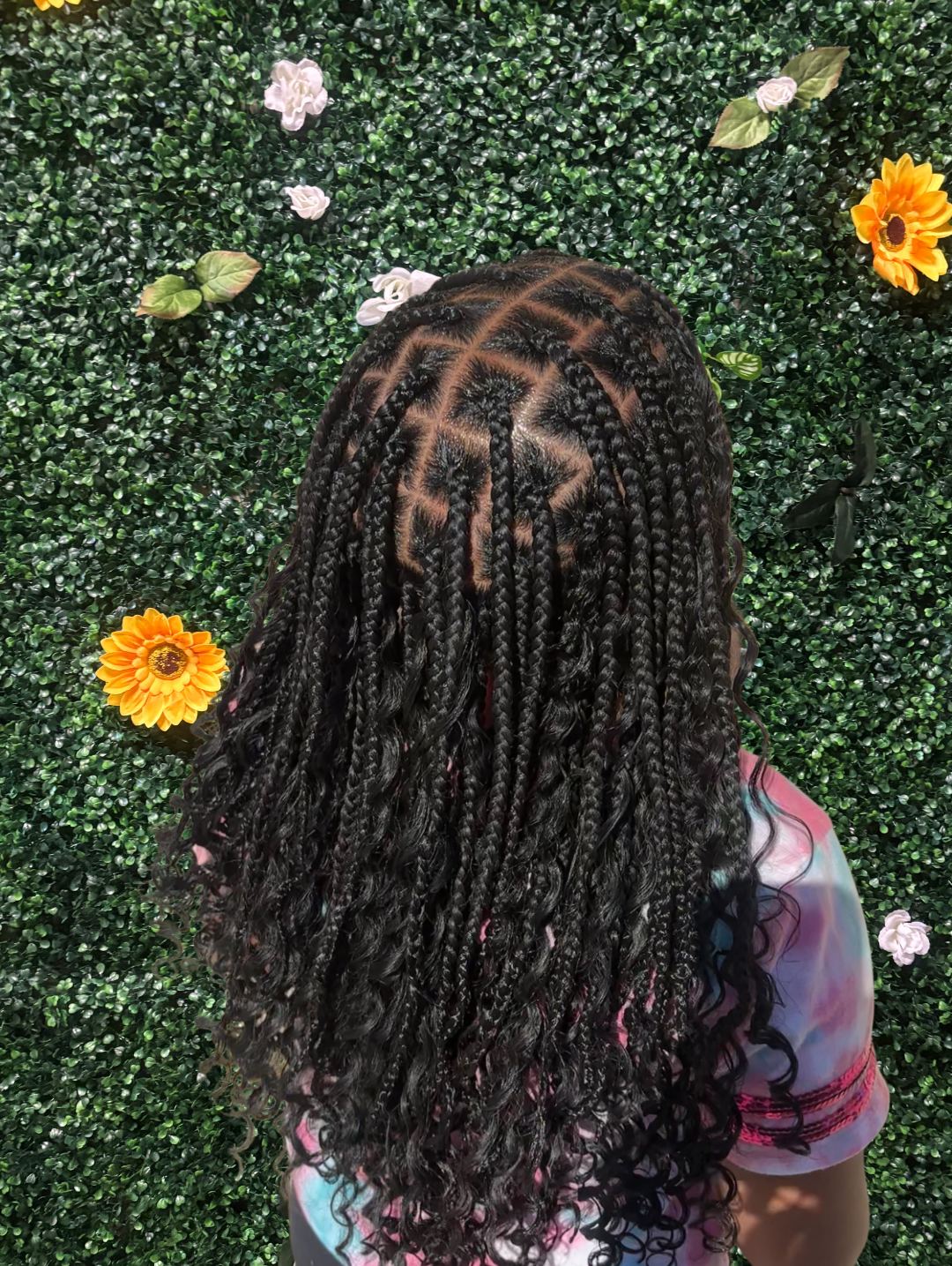 Back view of a person with long, black, knotless braids with curly ends, standing in front of a green faux hedge wall decorated with white, pink, and orange flowers.