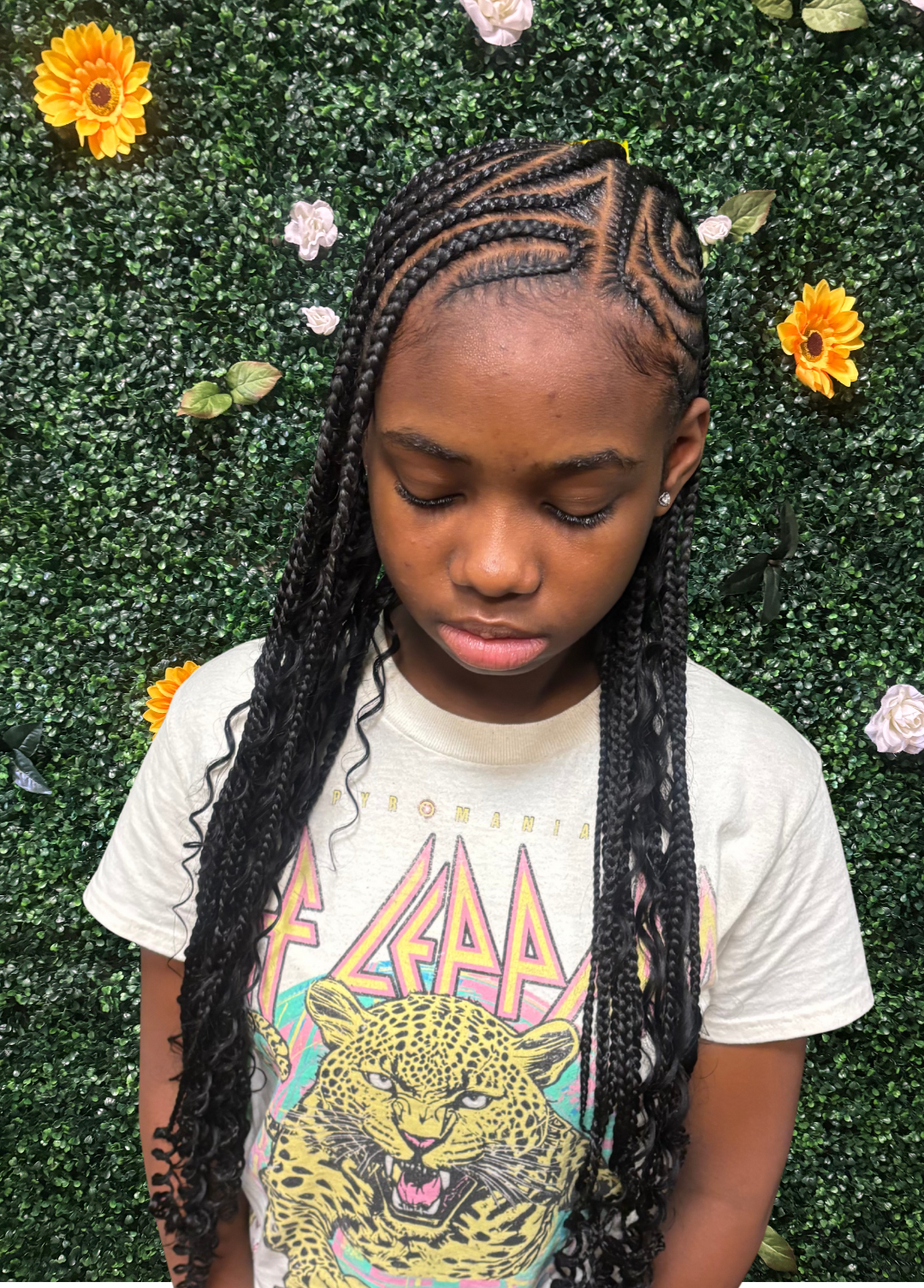 A girl with braided lemonade knotless braids and a graphic T-shirt standing in front of a green hedge wall with flowers.