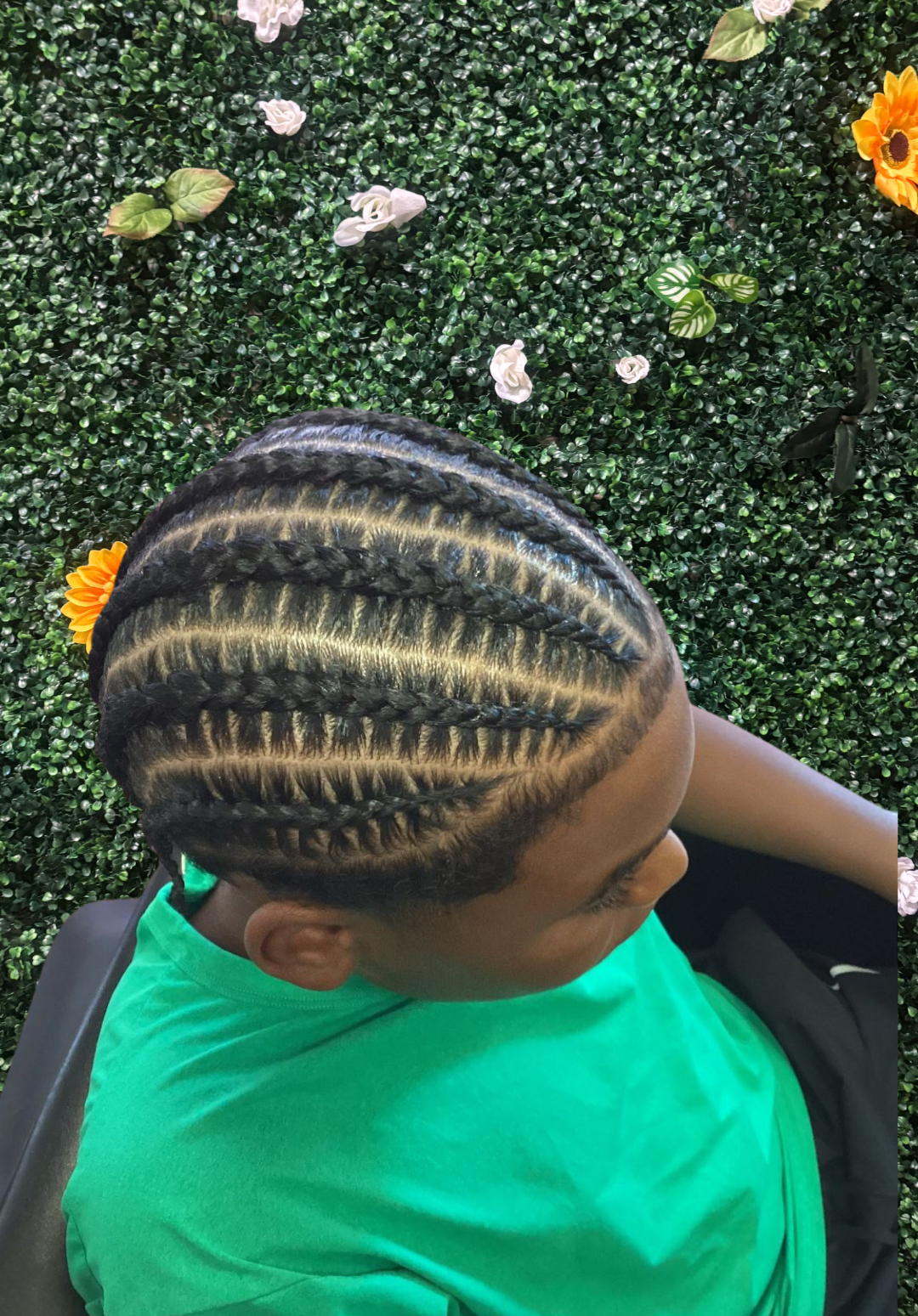 Top view of a person with cornrow braids and blonde highlights, sitting against a green artificial leafy wall with flowers.