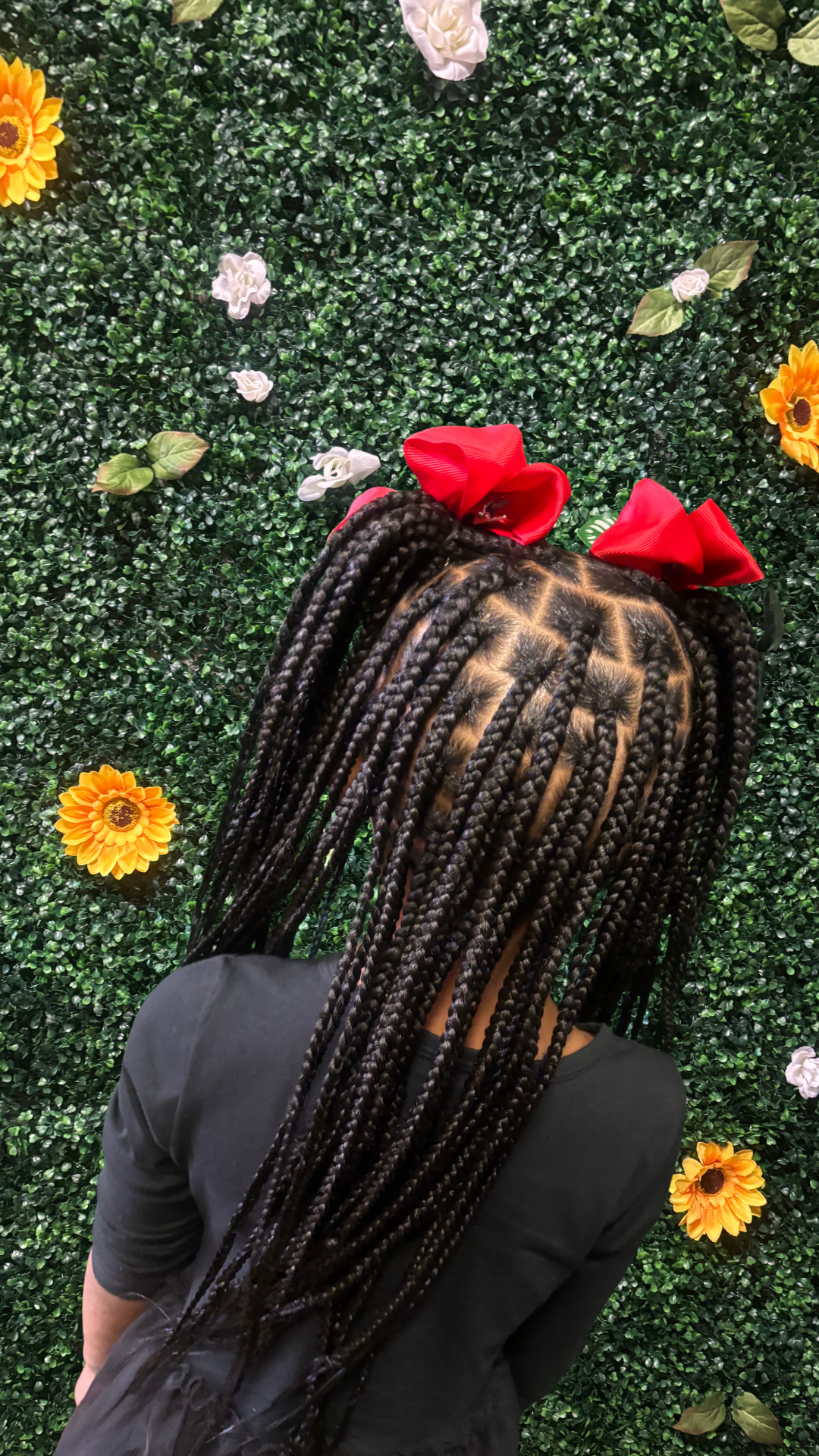 A person with long traditional box braids standing in front of a green floral wall decorated with sunflowers, white roses, and green leaves, wearing a black top and a red bow headband.
