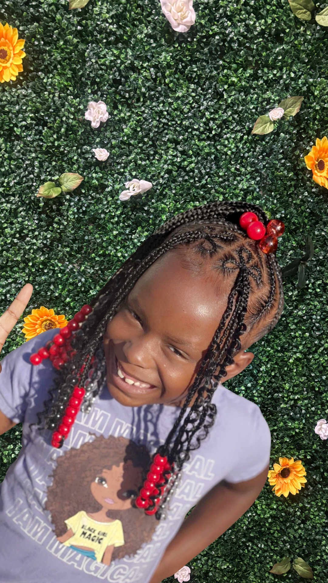 A young girl with a braided ponytail with bangs decorated with red beads, smiling and making a peace sign, standing in front of a green leafy background decorated with sunflowers and white flowers.