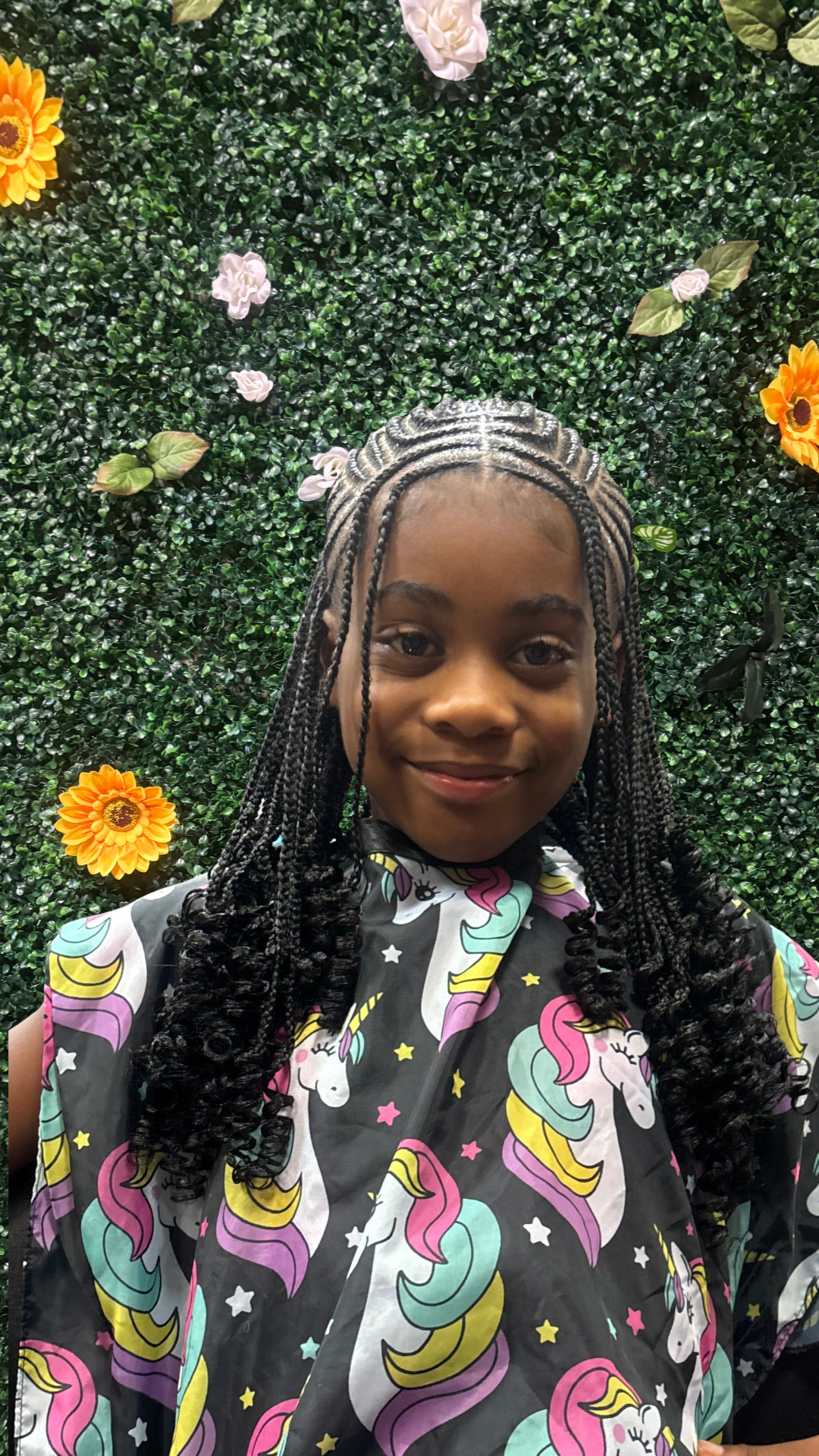 A young girl with braided hair styled in fulani knotless, smiling at the camera, standing in front of a green hedge wall decorated with white, purple, and yellow flowers, wearing a colorful unicorn-themed cape.