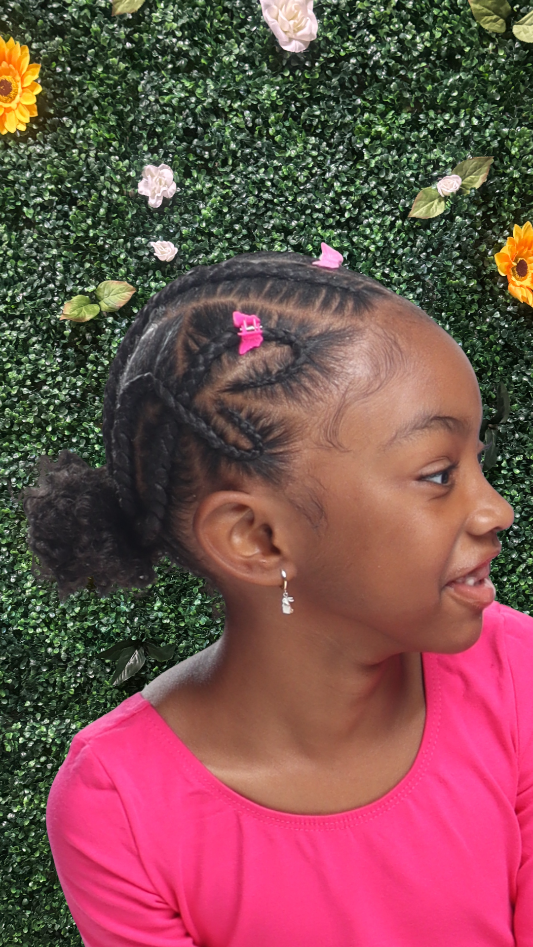 A young girl with stitch braided hairstyle decorated with pink hair clips, wearing a pink shirt, smiling, standing in front of a green leafy wall with colorful flowers.
