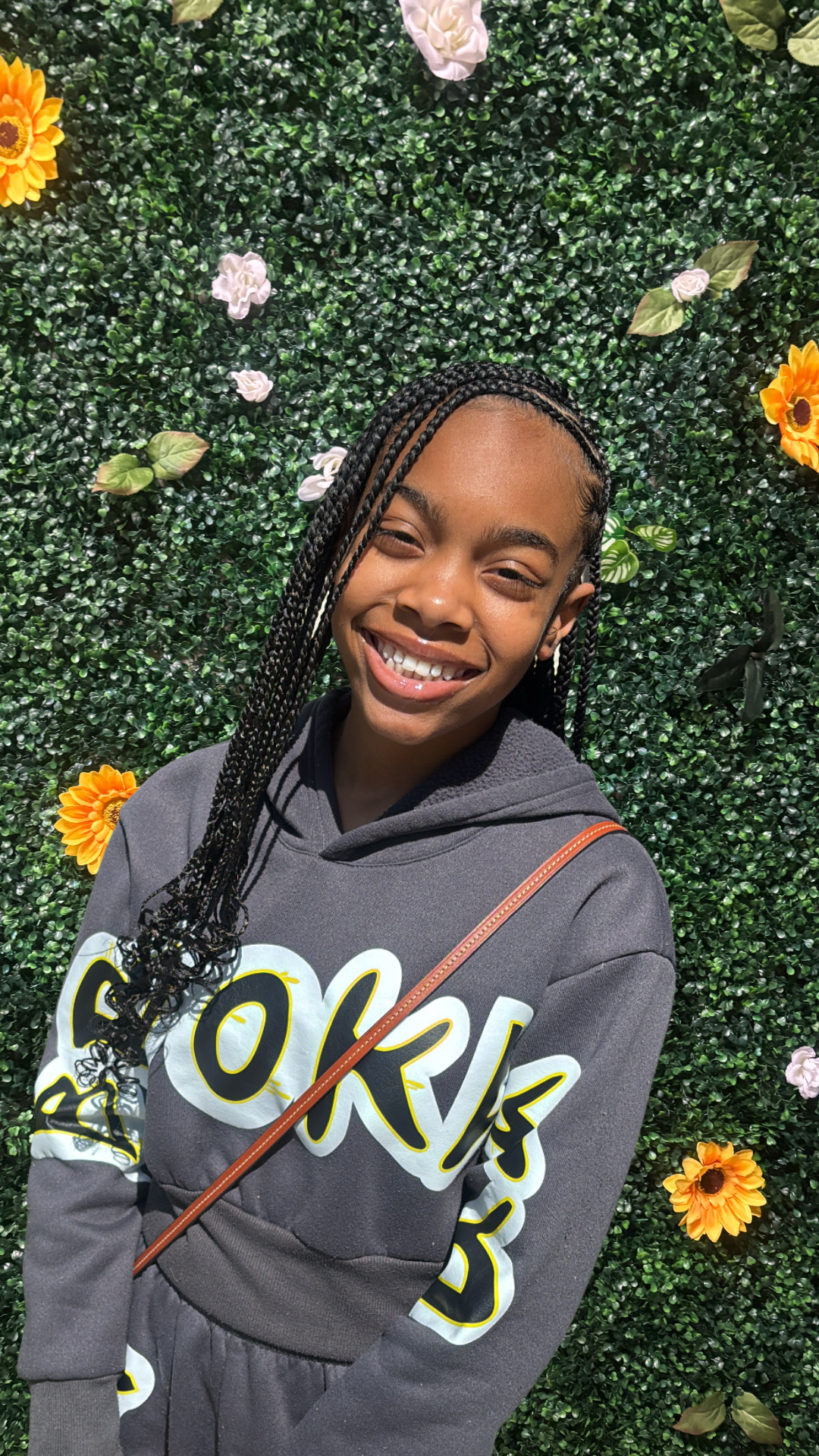 A young woman with braided hair smiling in front of a green leafy wall decorated with white, orange, and yellow flowers.