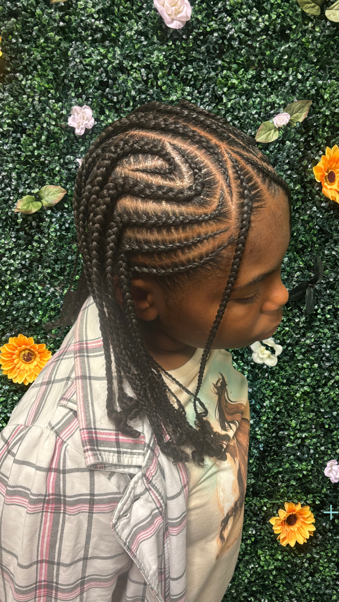 A young girl with braided hair, styled with cornrows and twists, standing against a green floral backdrop with white, yellow, and purple flowers.