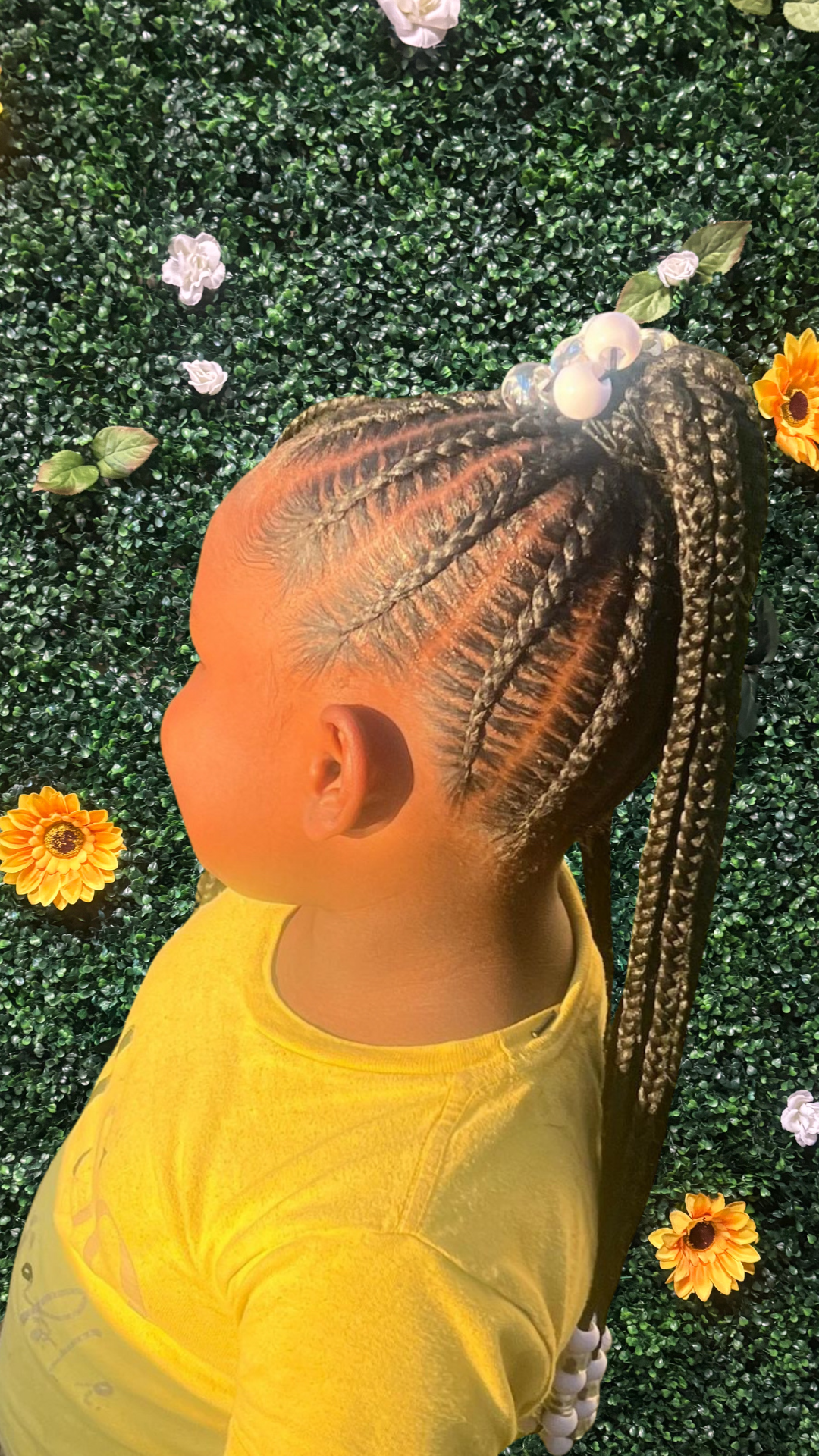 Side view of a young girl with a stitch braids styled into a ponytail, decorated with white beads, wearing a yellow shirt, standing against a green leafy wall with flowers.