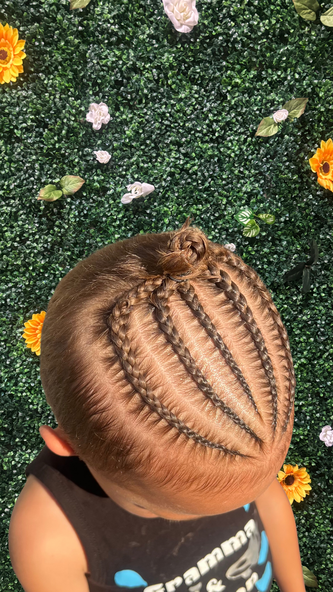 Close-up of a young child's head with stitch braided hair, placed against a green leafy backdrop decorated with white, yellow, and purple flowers.