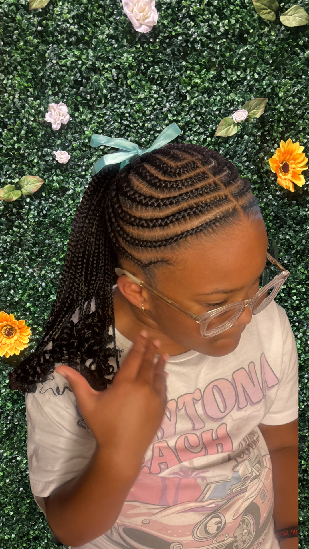 A child with braided middle-part ponytail and glasses shows off her hairstyle in front of a green floral background with white and yellow flowers.
