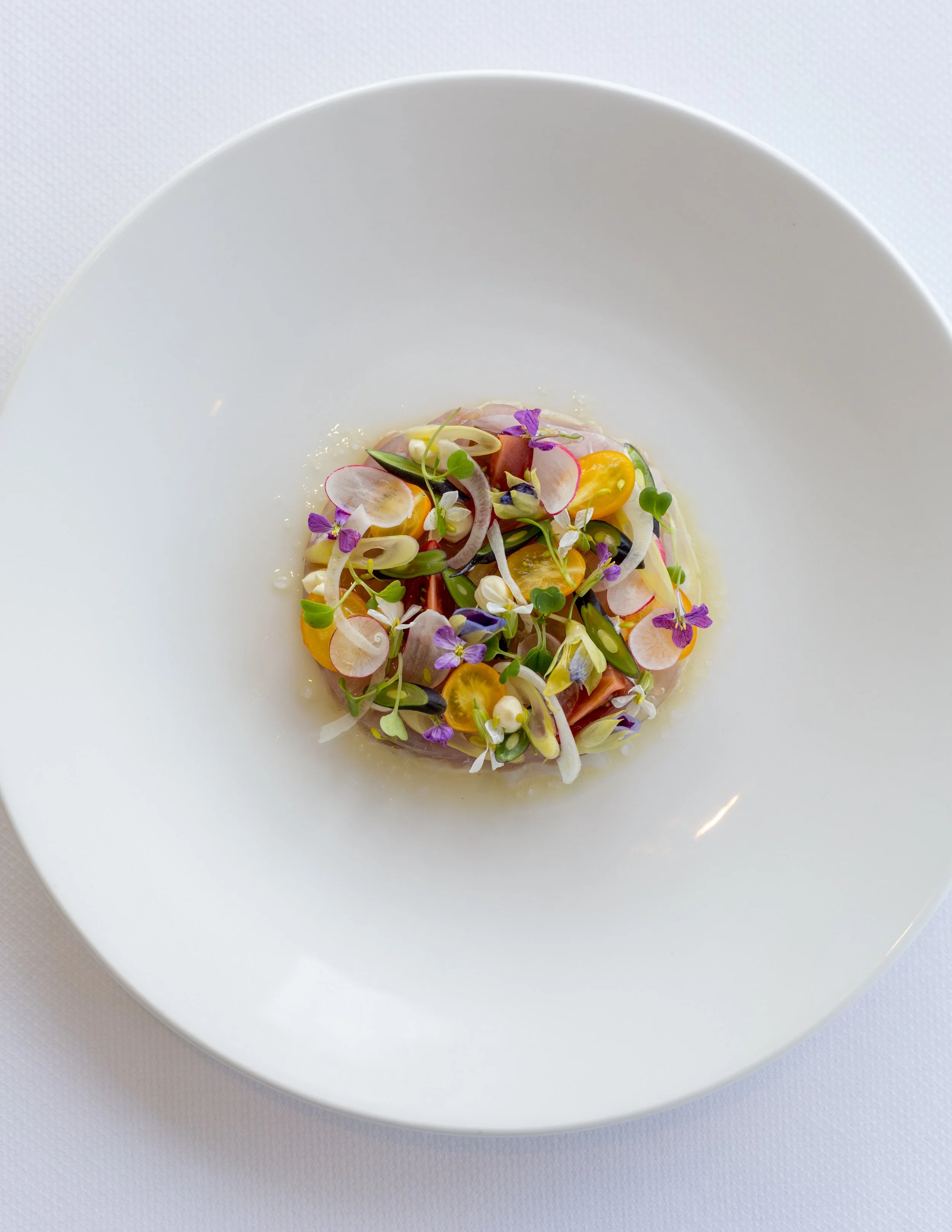 A white, elegant, round plate with a colorful, artistically arranged salad featuring sliced radishes, cherry tomatoes, microgreens, edible flowers, and herbs.