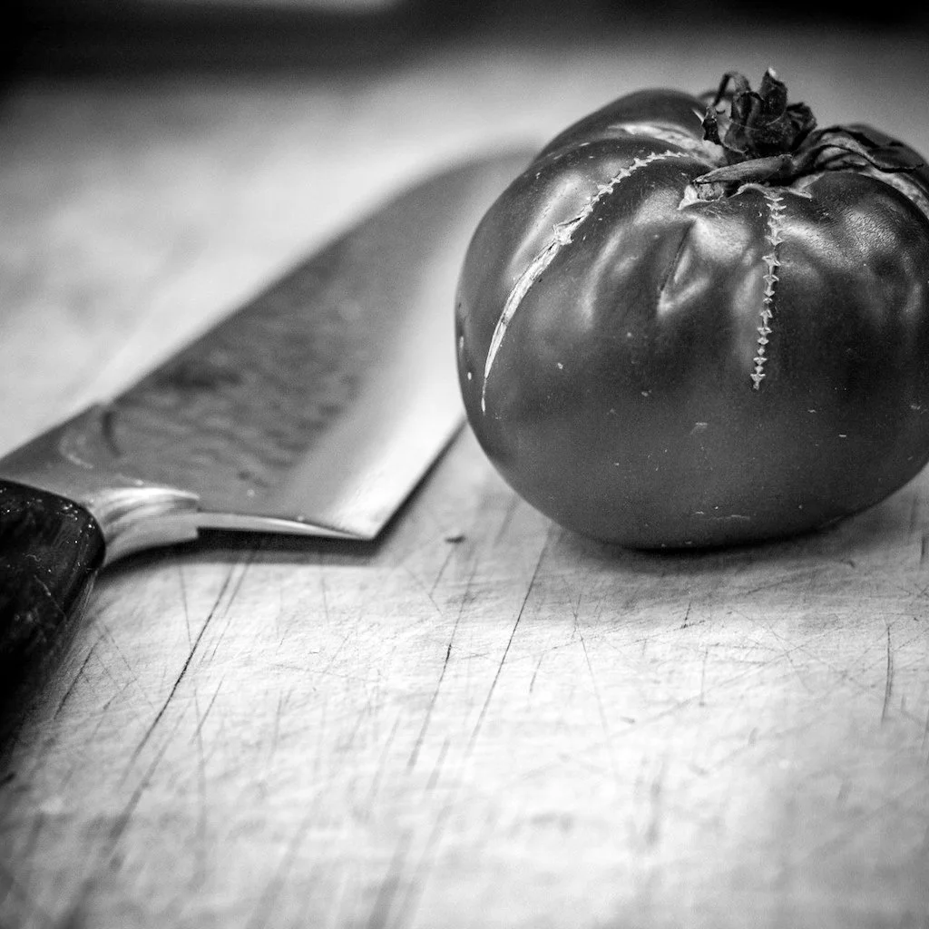 Black and white photo of a ripe tomato and a kitchen knife on a wooden cutting board.