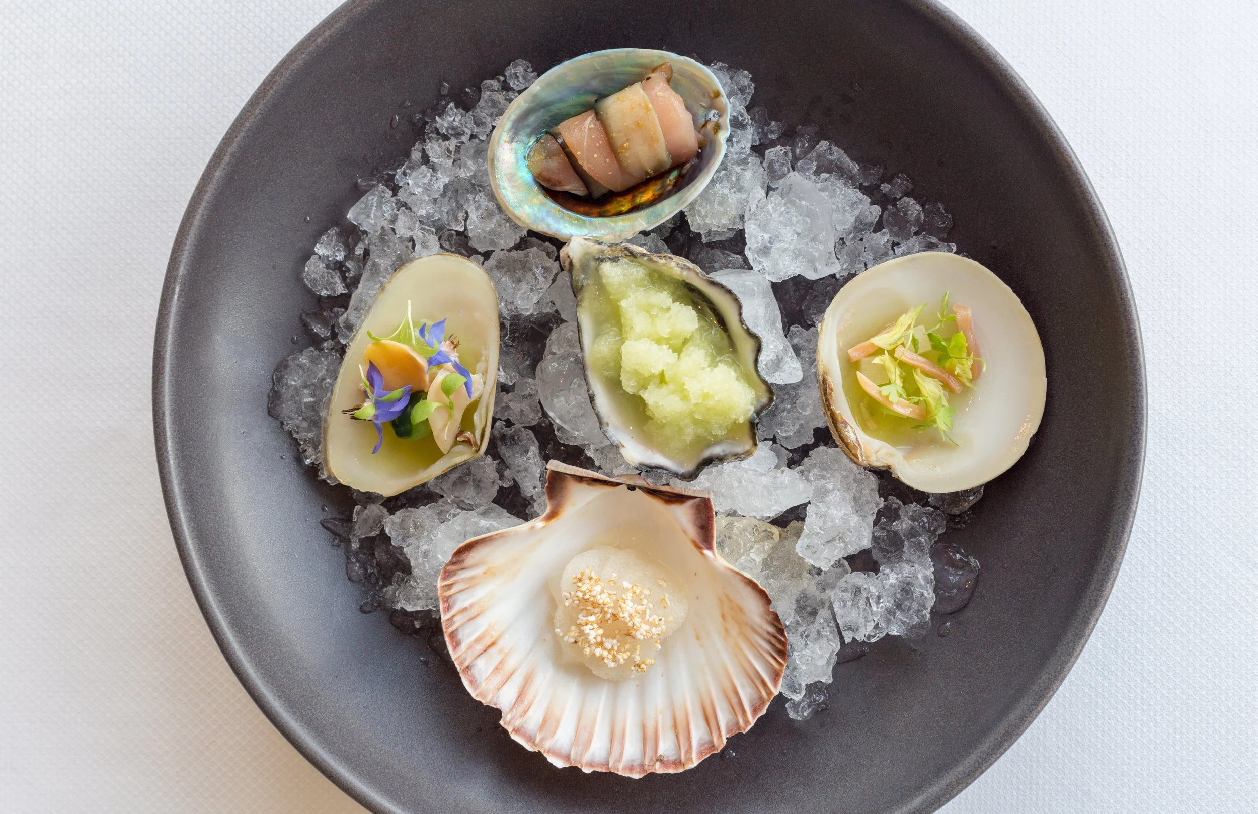A black plate with ice and five oysters, some garnished and others plain, arranged in a circle.