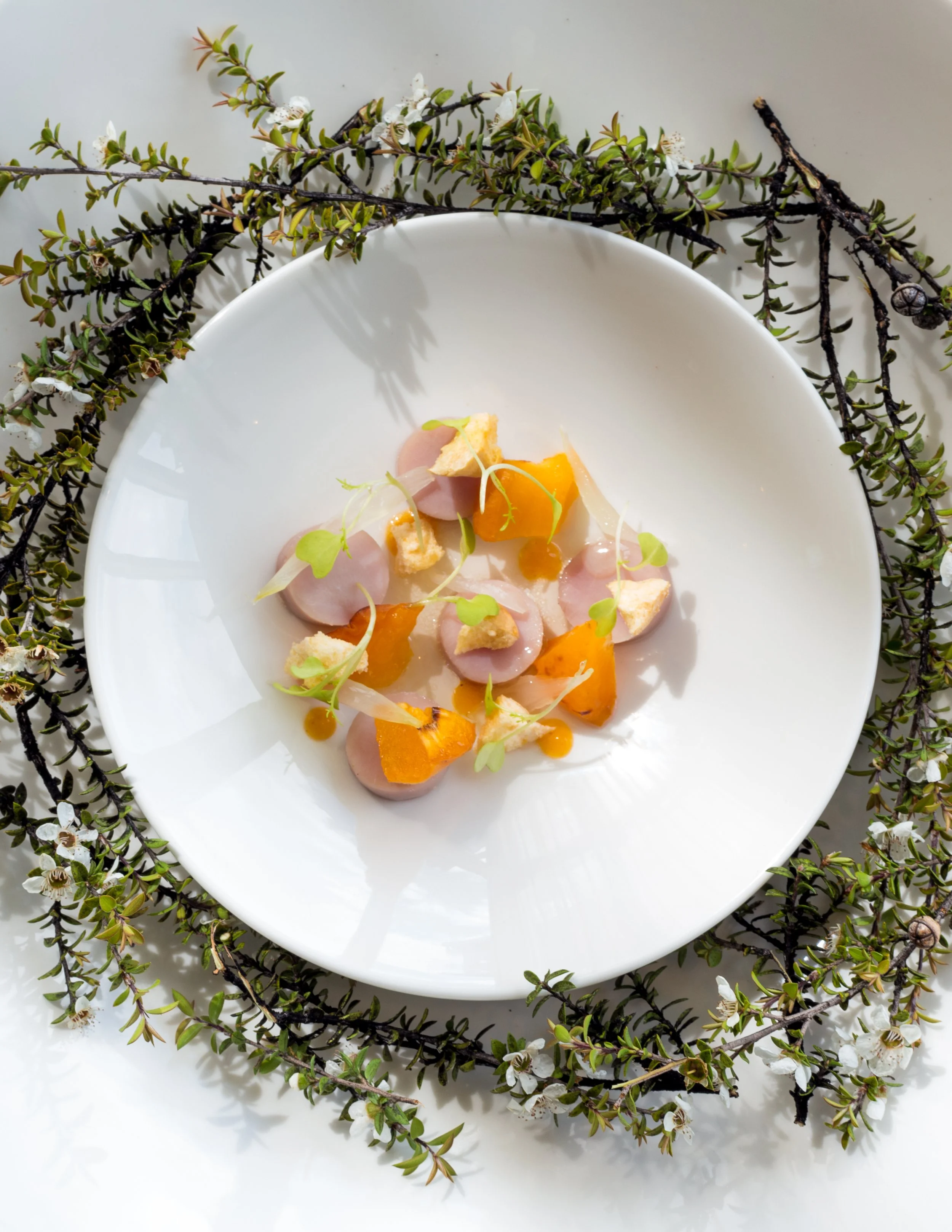 A white plate with a gourmet dish consisting of sliced meat, orange sauce, microgreens, and small crispy toppings, surrounded by sprigs of flowering greenery on a light surface.