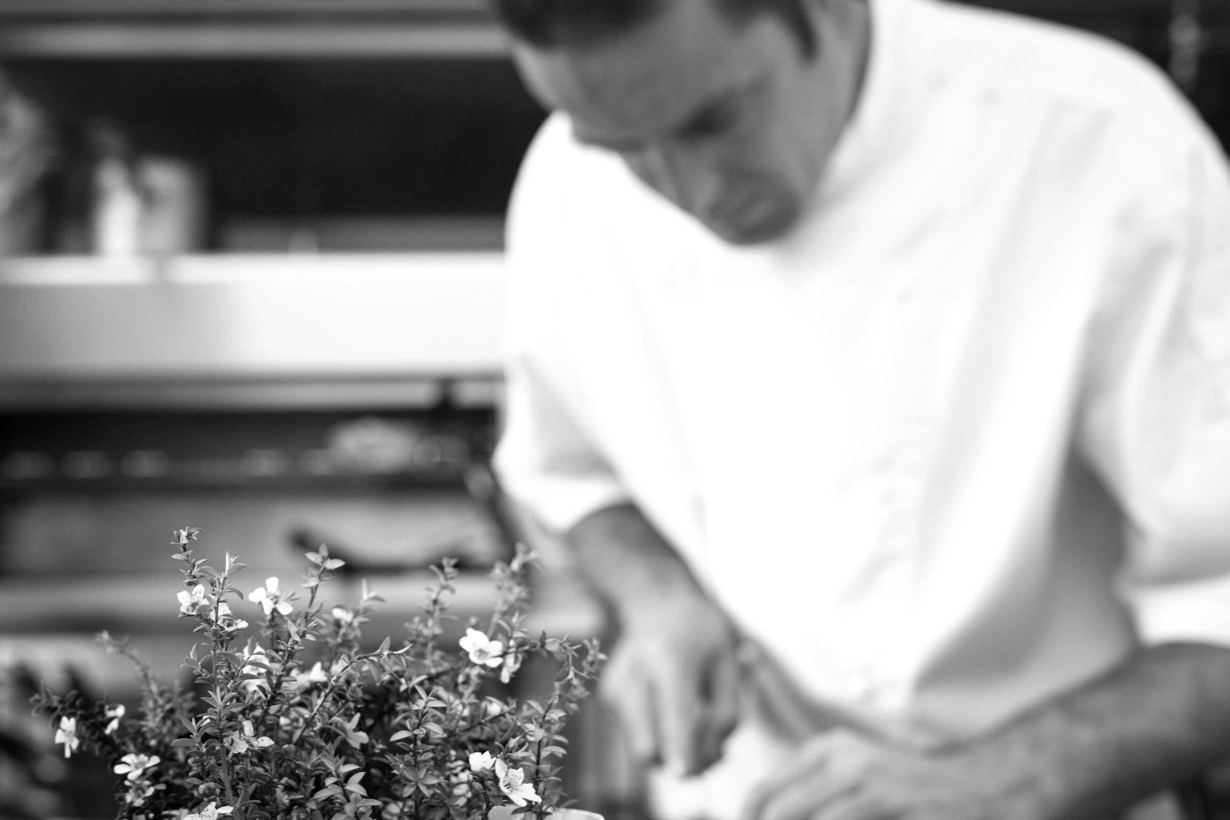 A chef with short hair leaning over a table preparing food, focused on work in a kitchen.