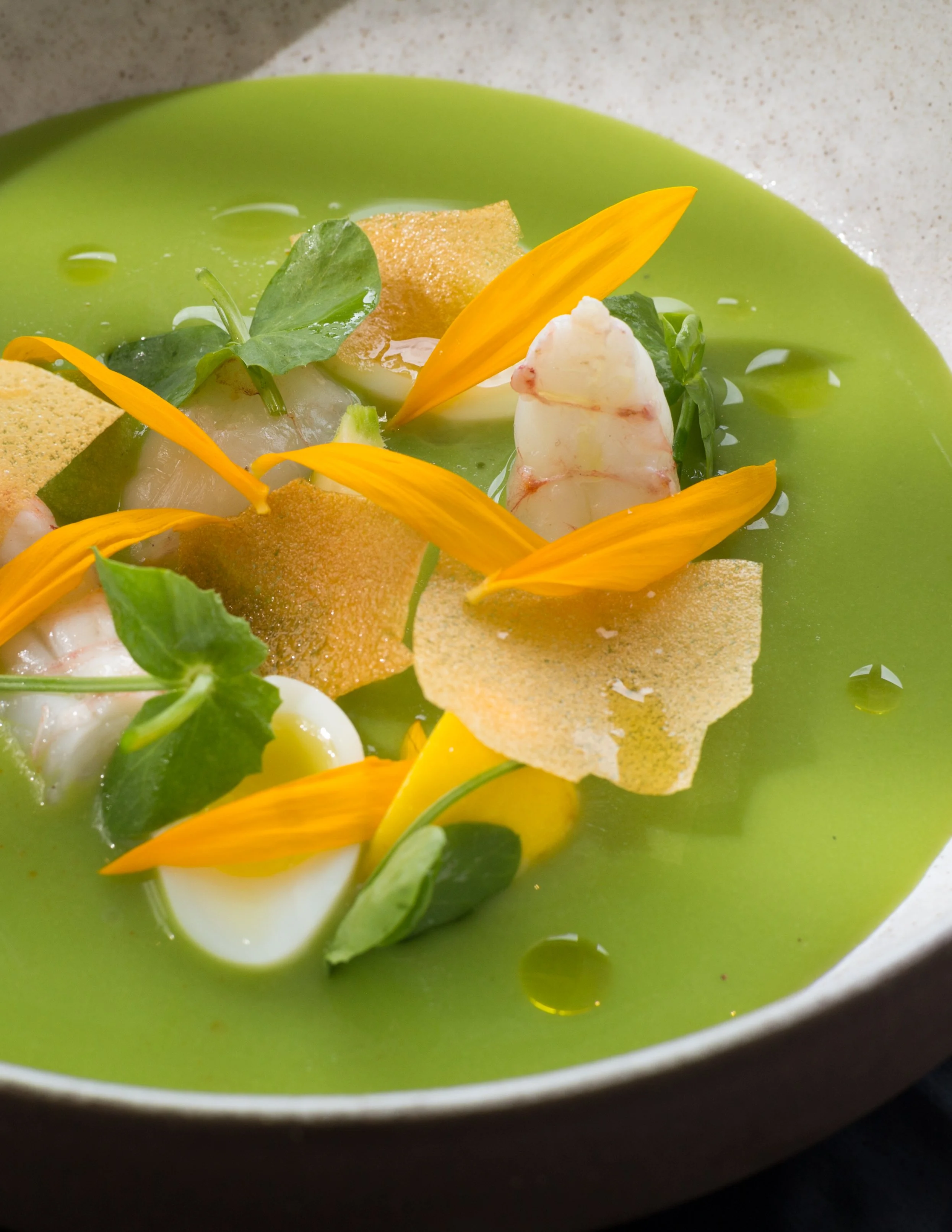 Close-up of a gourmet dish with green broth, garnished with edible flowers, microgreens, crispy chips, and a halved boiled egg in a green bowl.