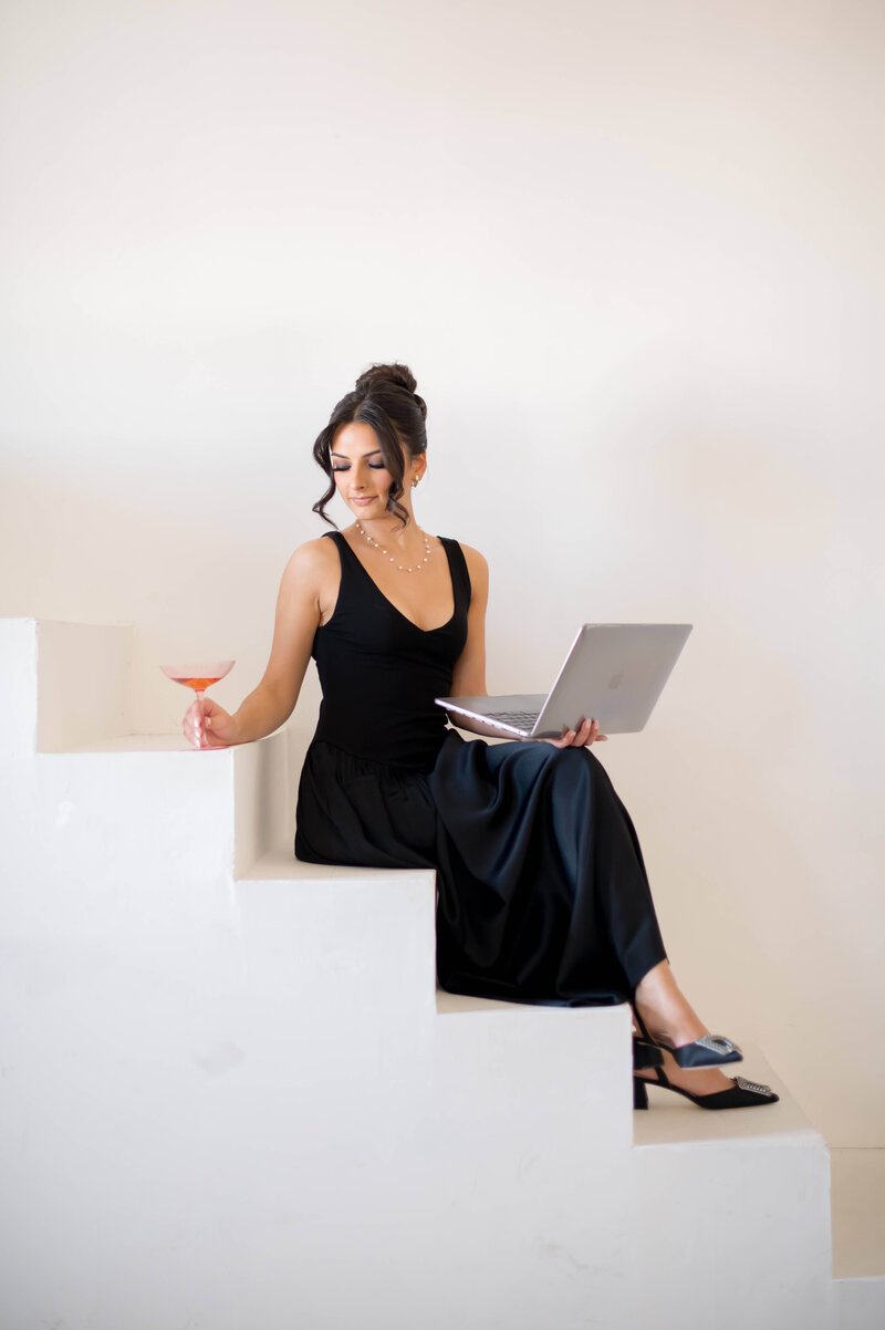 A woman in a black dress sitting on white stairs, holding a laptop in her lap and a cocktail in her hand, looking down with a slight smile.