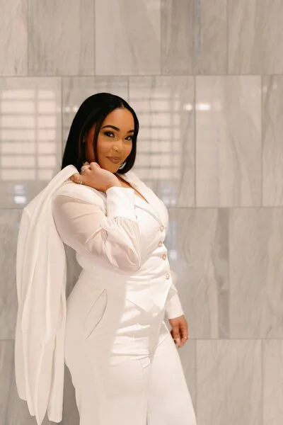 A woman in white clothing standing in front of a gray tiled wall, posing with one hand on her shoulder and holding a white jacket over her shoulder.