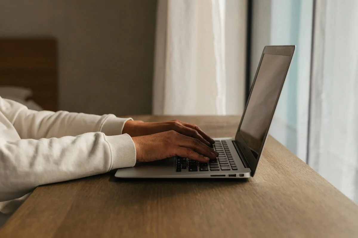 Person wearing a white sweatshirt typing on a silver laptop at a wooden desk in a room with curtains.