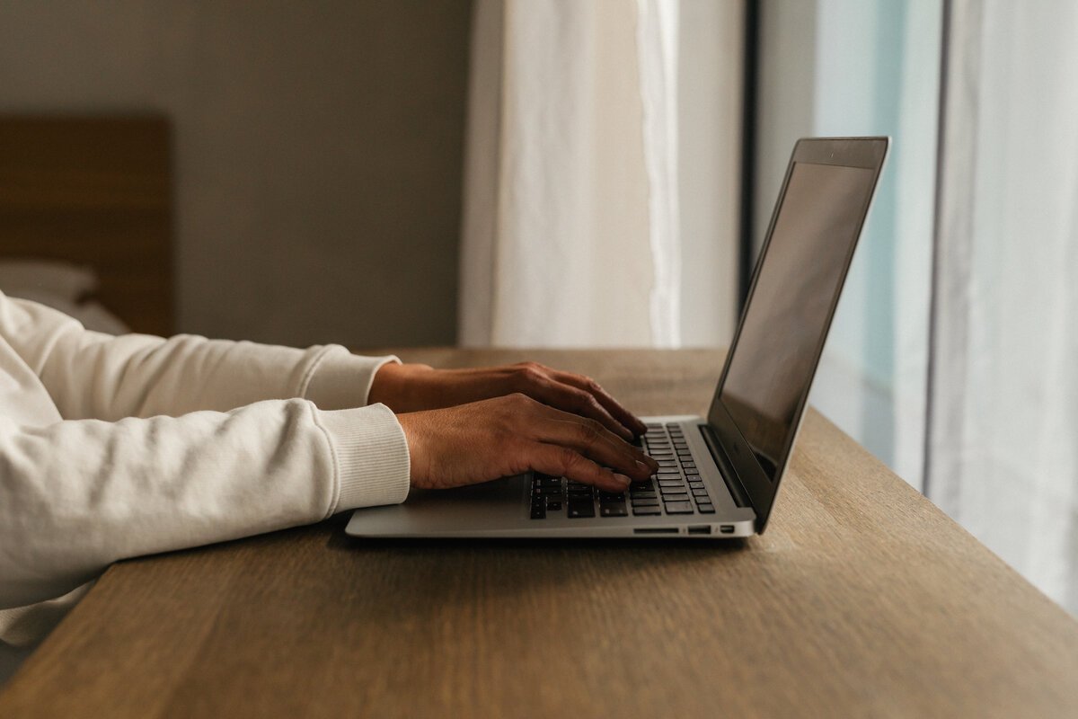 Person wearing a white long-sleeve shirt typing on a laptop at a wooden table near a window with curtains.