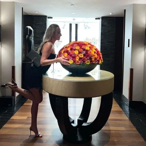 A woman with blonde hair, wearing a black dress and high heels, stands next to a large flower arrangement on a round table in a modern indoor space. The arrangement consists of a sphere of colorful roses on top of a unique black and beige table.