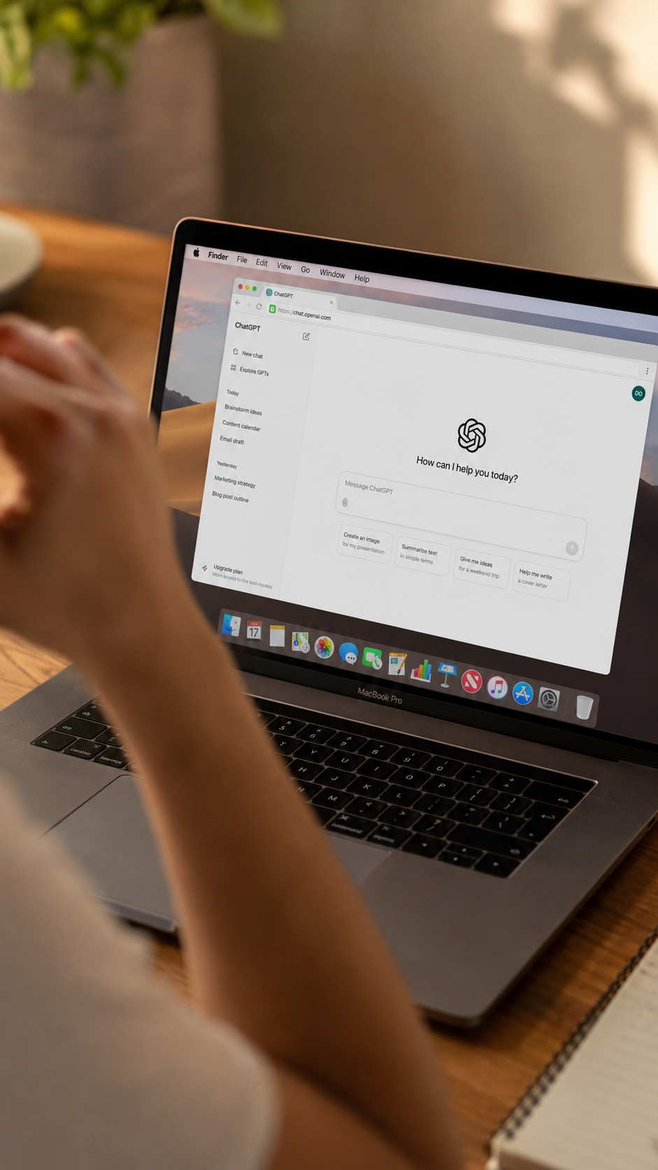 A person sitting at a wooden desk using a MacBook Pro laptop with the ChatGPT website open on the screen. The person has their hand near their face, and appearing to think or concentrate. There is a notebook with a pen near the laptop and some greenery in the background.