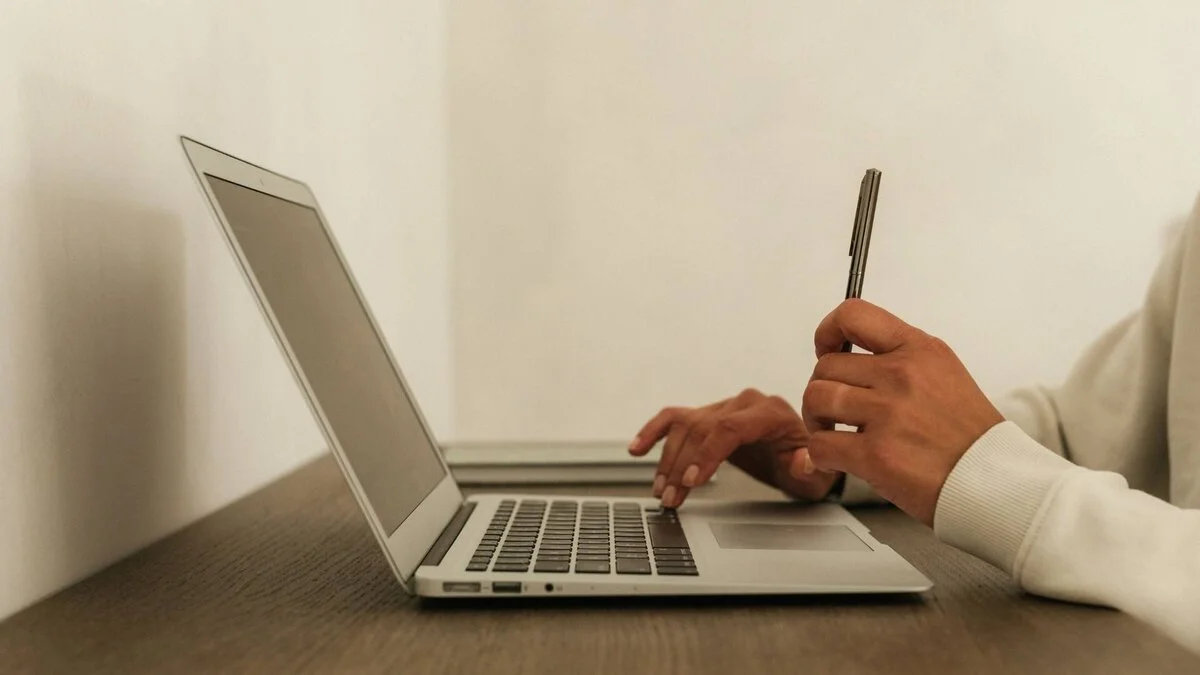 Person using a laptop computer and holding a pen while seated at a wooden desk.