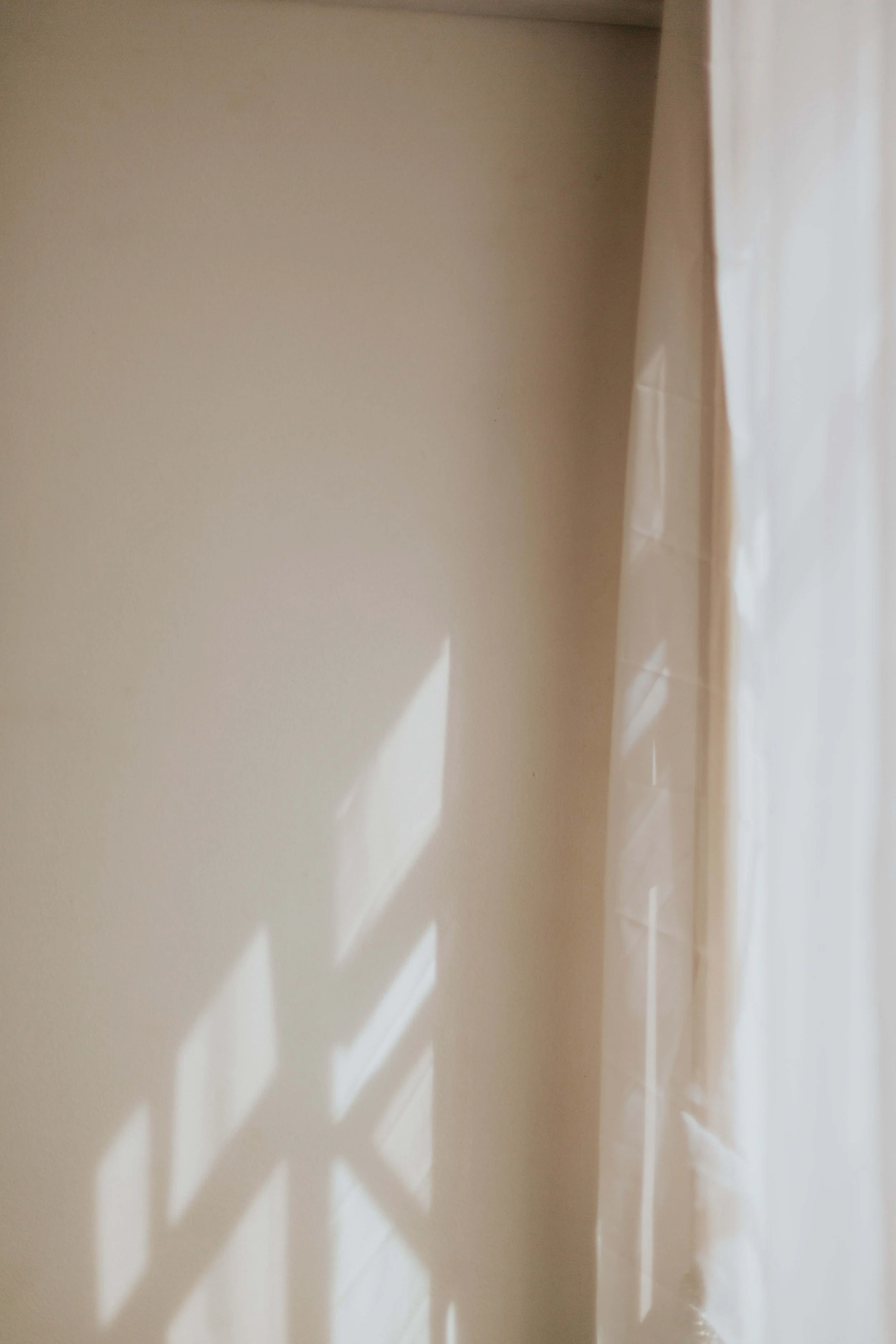 A beige wall with shadows cast by window blinds and a sheer curtain on the right side.
