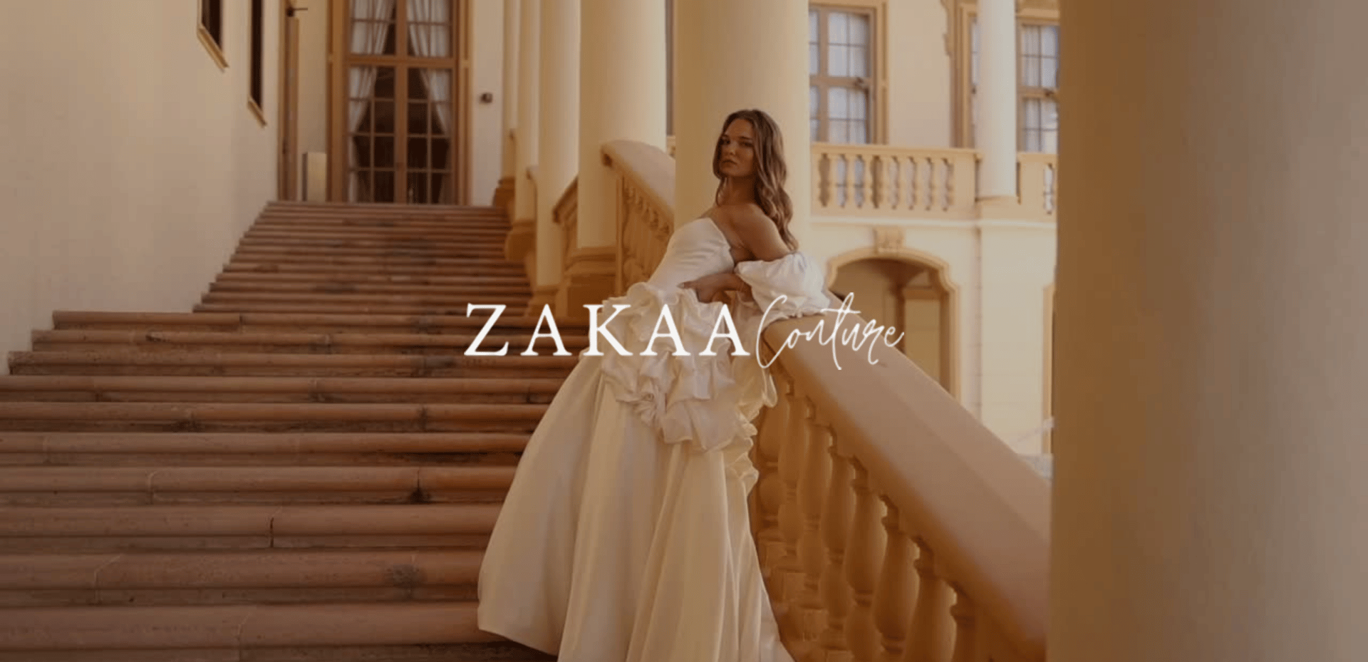 A woman in a cream-colored wedding gown standing on a grand staircase inside a historic building with large windows and columns. The text 'ZAKAA Couture' is overlaid on the image.