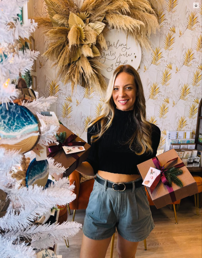 A woman smiling in a cozy decorated room, holding a wrapped gift box with a ribbon and Christmas-themed decorations, standing near a white Christmas tree and holiday decor.