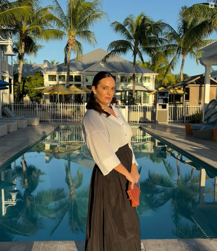 A woman stands by a swimming pool at a tropical resort with palm trees, surrounding lounge chairs, and a building with a glass facade in the background during the daytime.