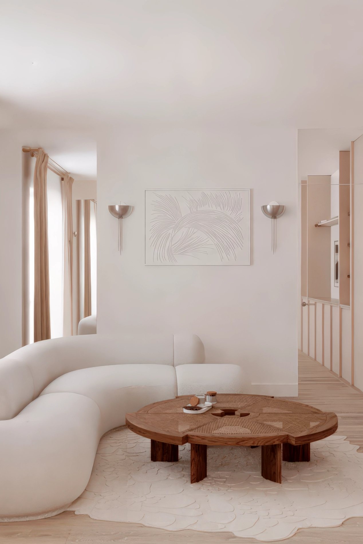 Intérieur d'un salon avec canapé blanc en forme de U, table en bois rond, mur blanc avec décoration de feuille, lampes murales, et fenêtres avec rideaux couleur crème.